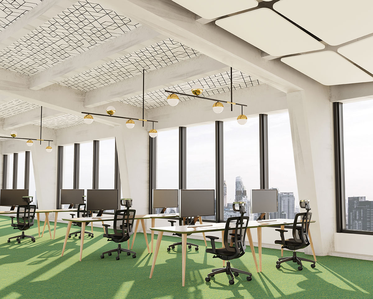 A modern office space with large windows offering a city view. The room has green flooring, multiple workstations with computers, black office chairs, and contemporary lighting. The ceiling features a unique design with printed acoustic felt ceiling panels and exposed beams in a light grid pattern.