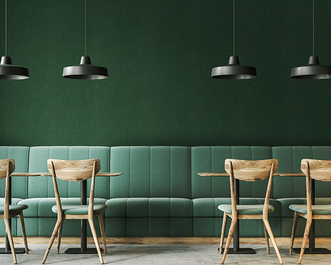 A modern dining area features a long, green upholstered bench along a wall adorned with matching acoustic felt wall panels. Wooden chairs and tables are arranged in front of the bench. Three black pendant lights hang from the ceiling, contributing to the minimalist aesthetic.