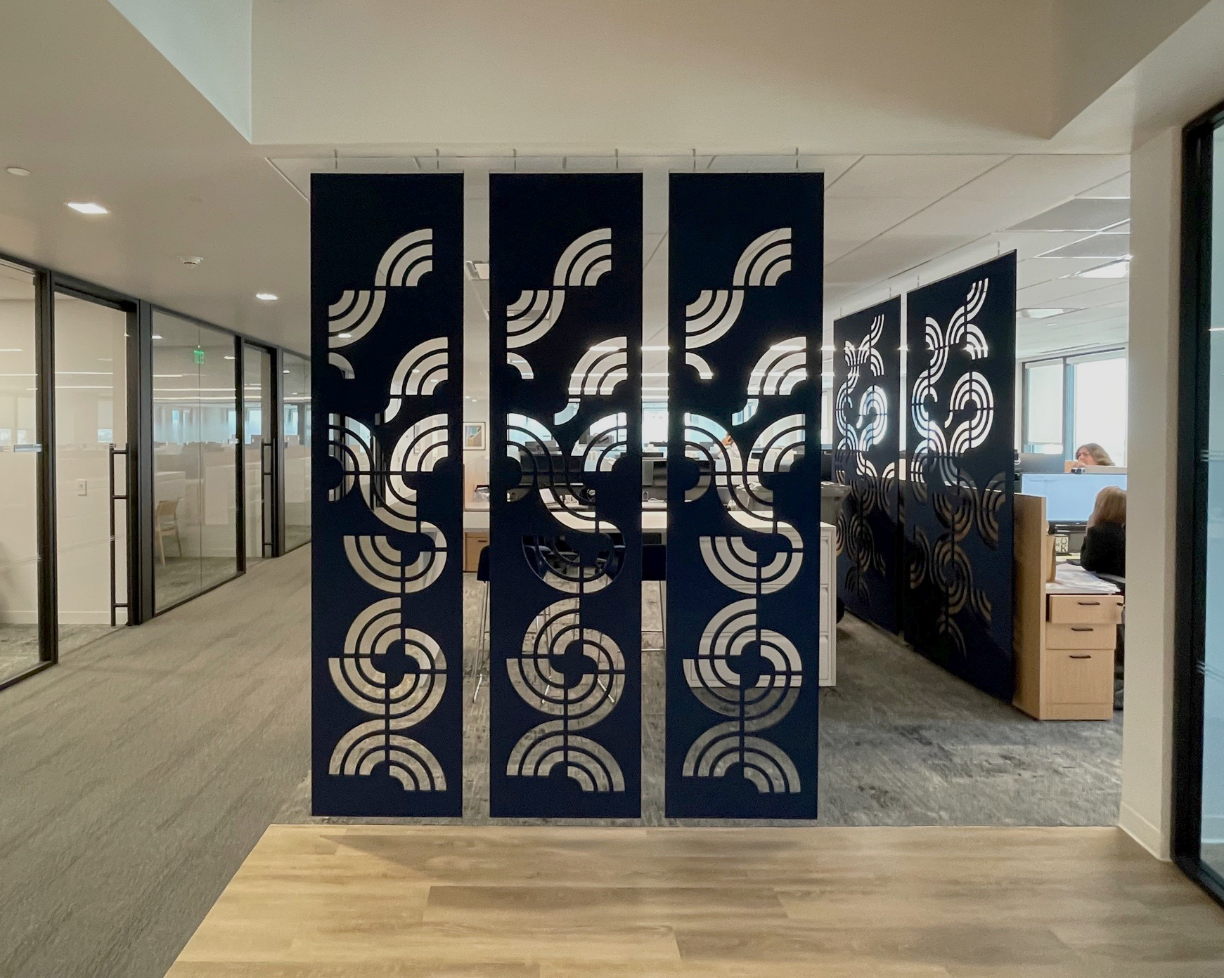 Modern office space with patterned dark acoustic felt screens acting as room dividers. The screens feature a geometric design with contrasting light shapes. Glass-walled offices and workstations are visible in the background, with grey carpeting and wooden flooring in the foreground.