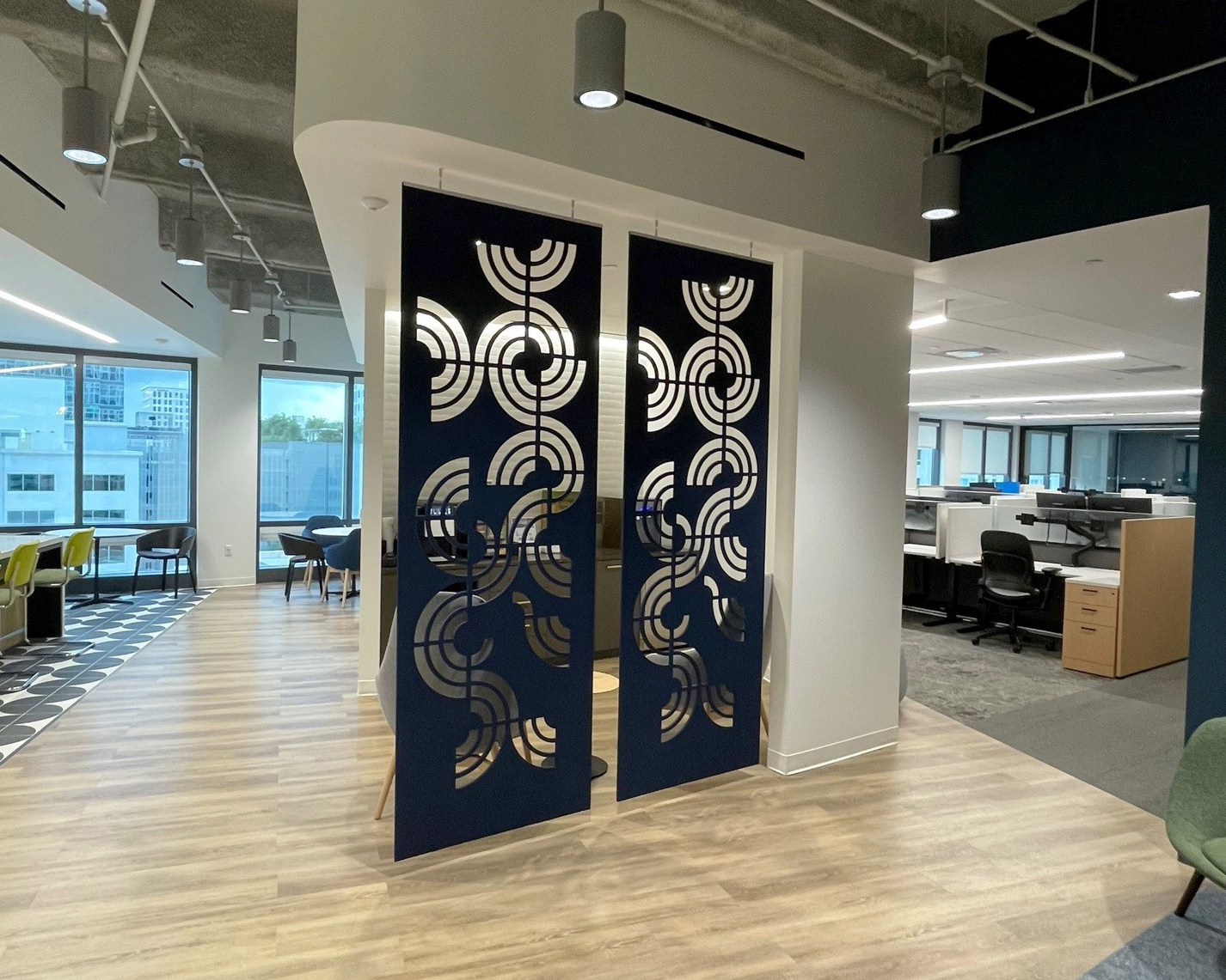 Modern office space with wooden flooring, open workstations, and geometric-patterned partitions. The ceiling is exposed, and there are large windows allowing natural light inside. Acoustic felt screens with circular cutouts divide the area.
