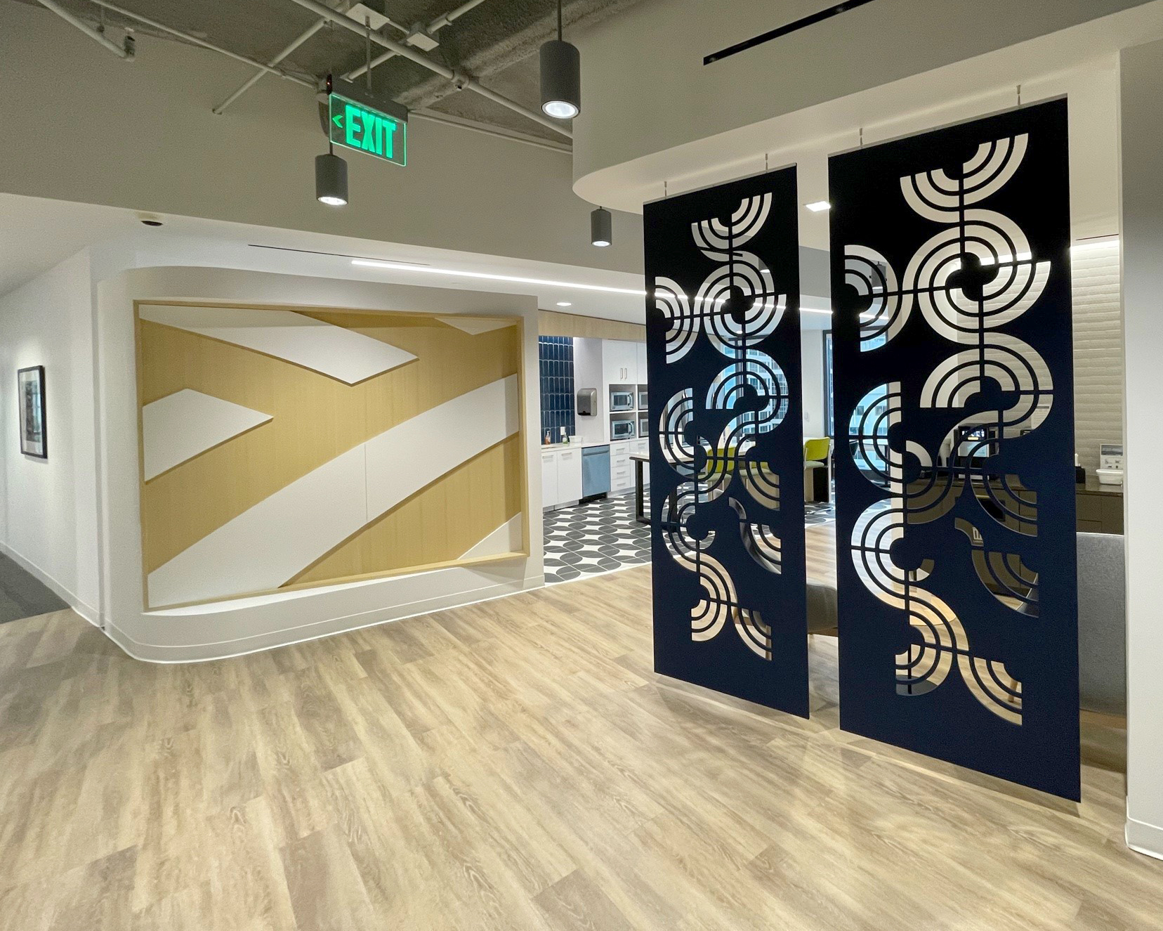 Modern office space with wooden floors, white walls, and a geometric art installation on the left wall. A section of the wall features laser-cut acoustic felt wall screens with circular patterns, partially separating the area. In the background is a kitchen area with a checkerboard floor.
