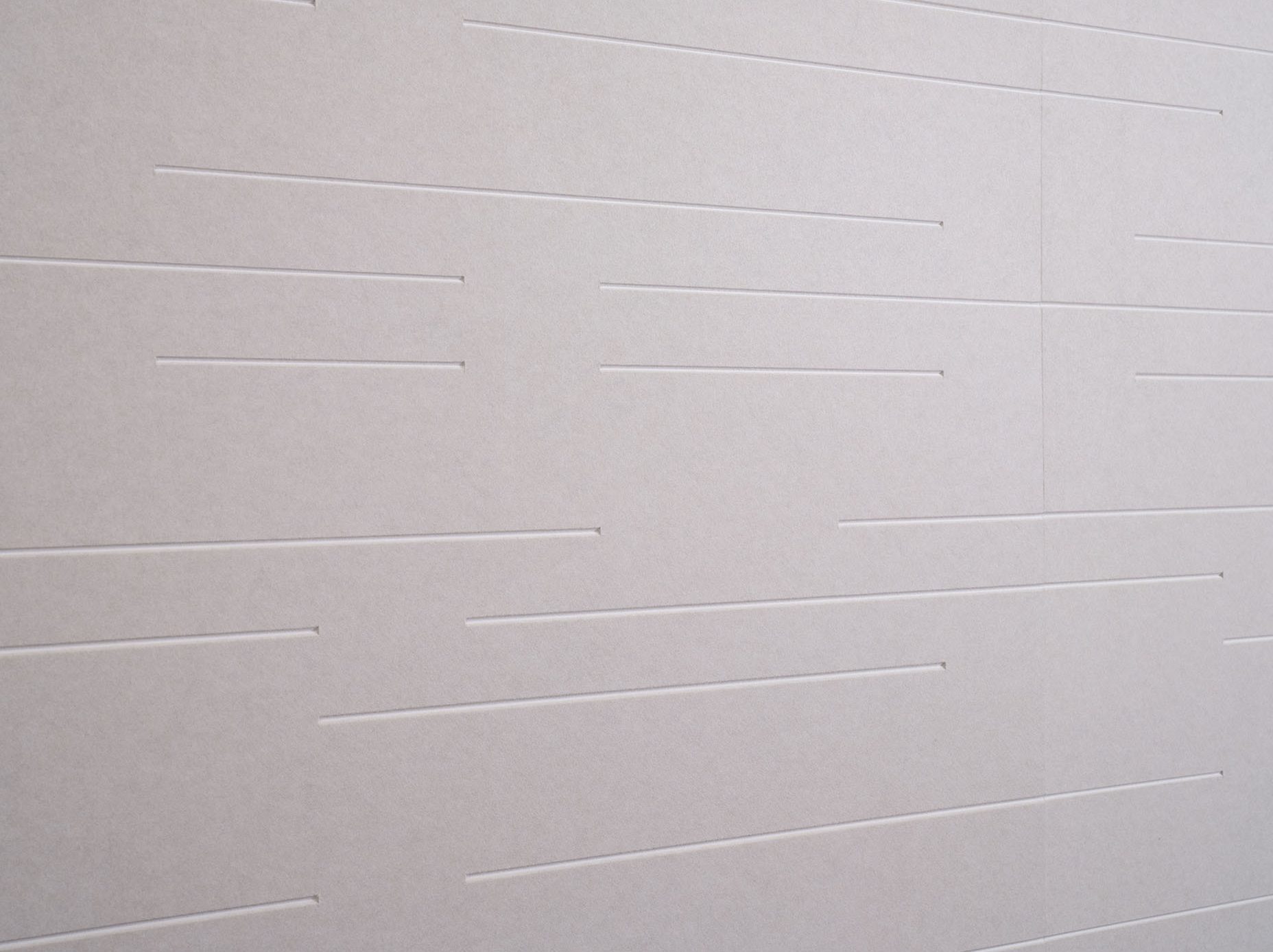 A close-up view of a beige wall with narrow, horizontal indented lines arranged in a seemingly random pattern across the surface. The lines are thin and evenly spaced, creating a subtle, textured appearance on the otherwise plain background.