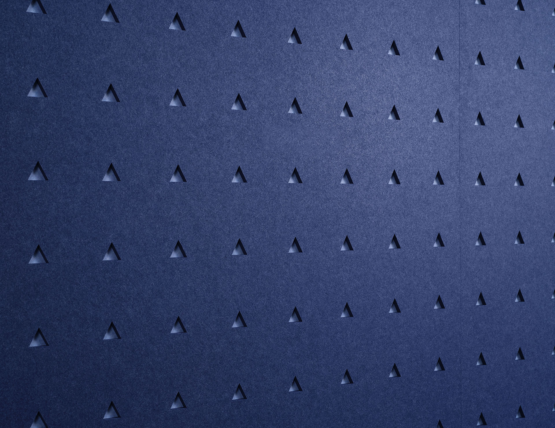 A dark blue textured wall with a repeating pattern of small, evenly spaced, downward triangular indentations. The light source casts subtle shadows, enhancing the geometric design and giving it a three-dimensional appearance.