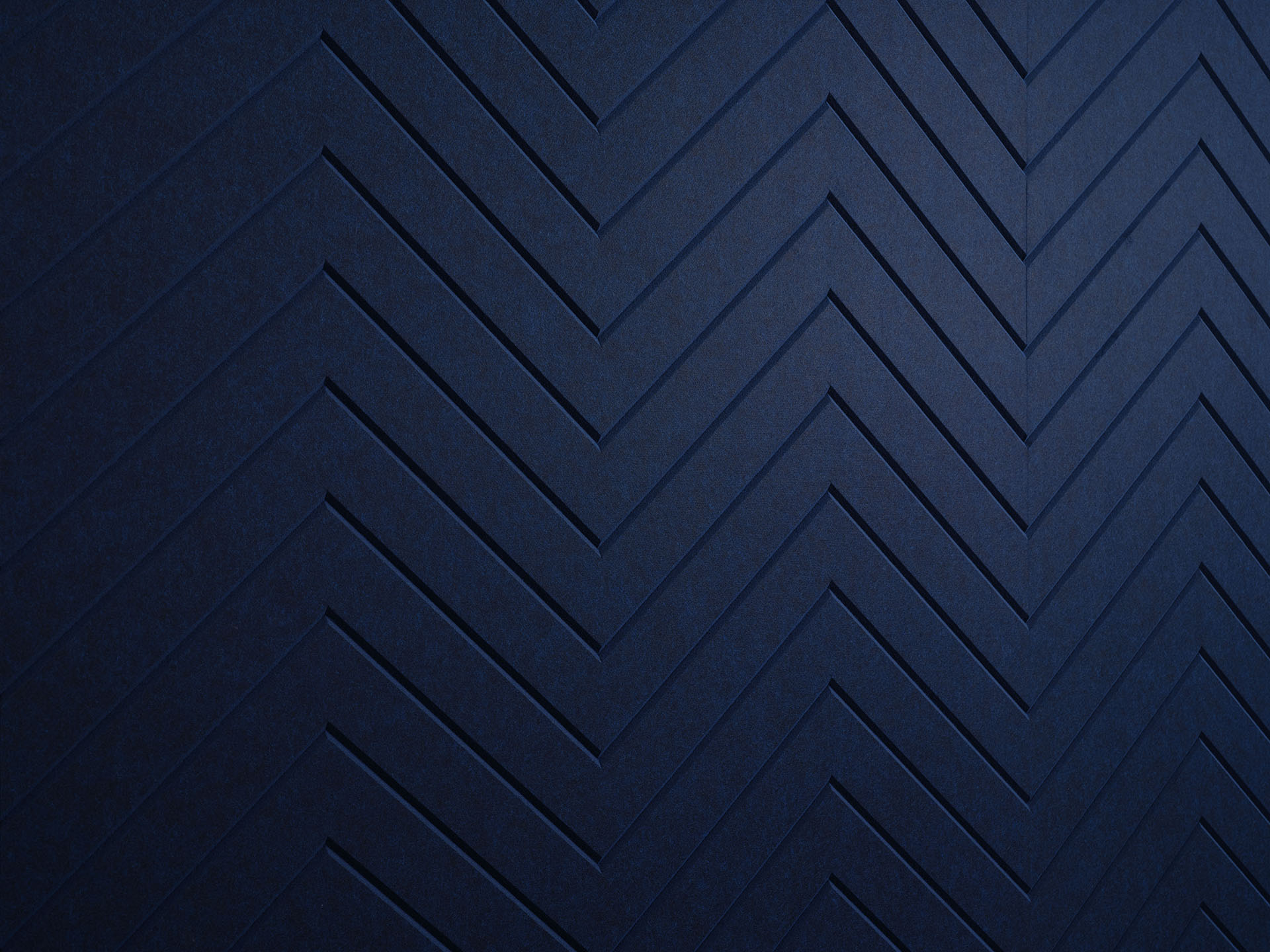 A close-up view of a textured acoustic felt panel with a dark blue herringbone pattern. The pattern consists of a series of zigzag lines creating a visually appealing and rhythmic design. The lighting accentuates the depth and contours of the texture.