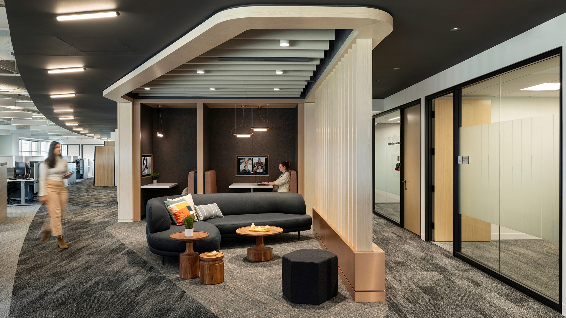 Modern office with sleek design features a small seating area with a black sofa, wooden tables, and hexagonal ottomans. Acoustic ceiling baffles enhance the space's acoustics while providing a stylish touch. A person works on a laptop at a booth in the background, with glass-walled conference rooms and workstations visible throughout the open space.