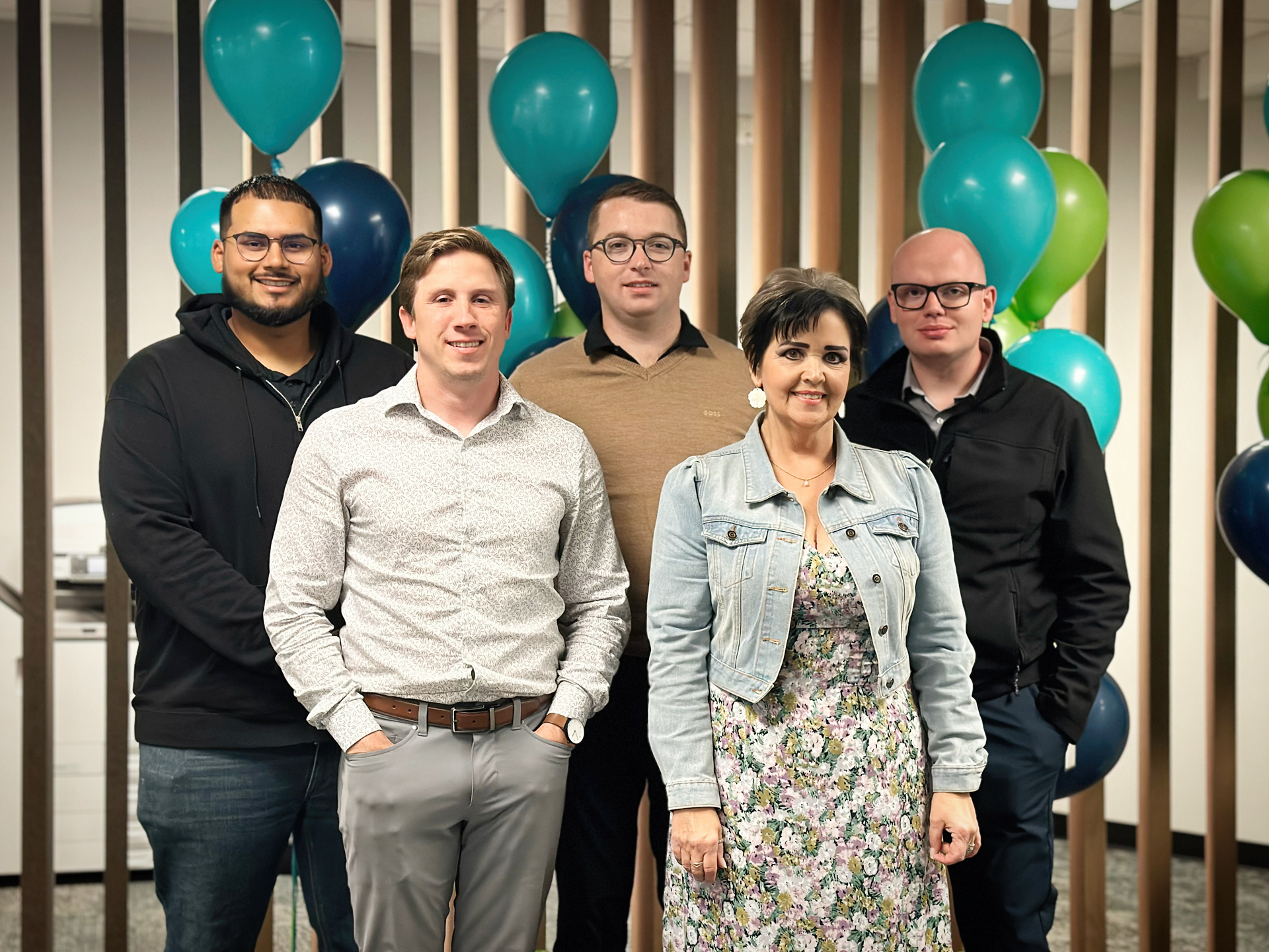 A group of five people standing in front of a wooden panel background with teal and blue balloons. Three men and one woman stand together, all smiling. They are dressed in casual and semi-casual attire.