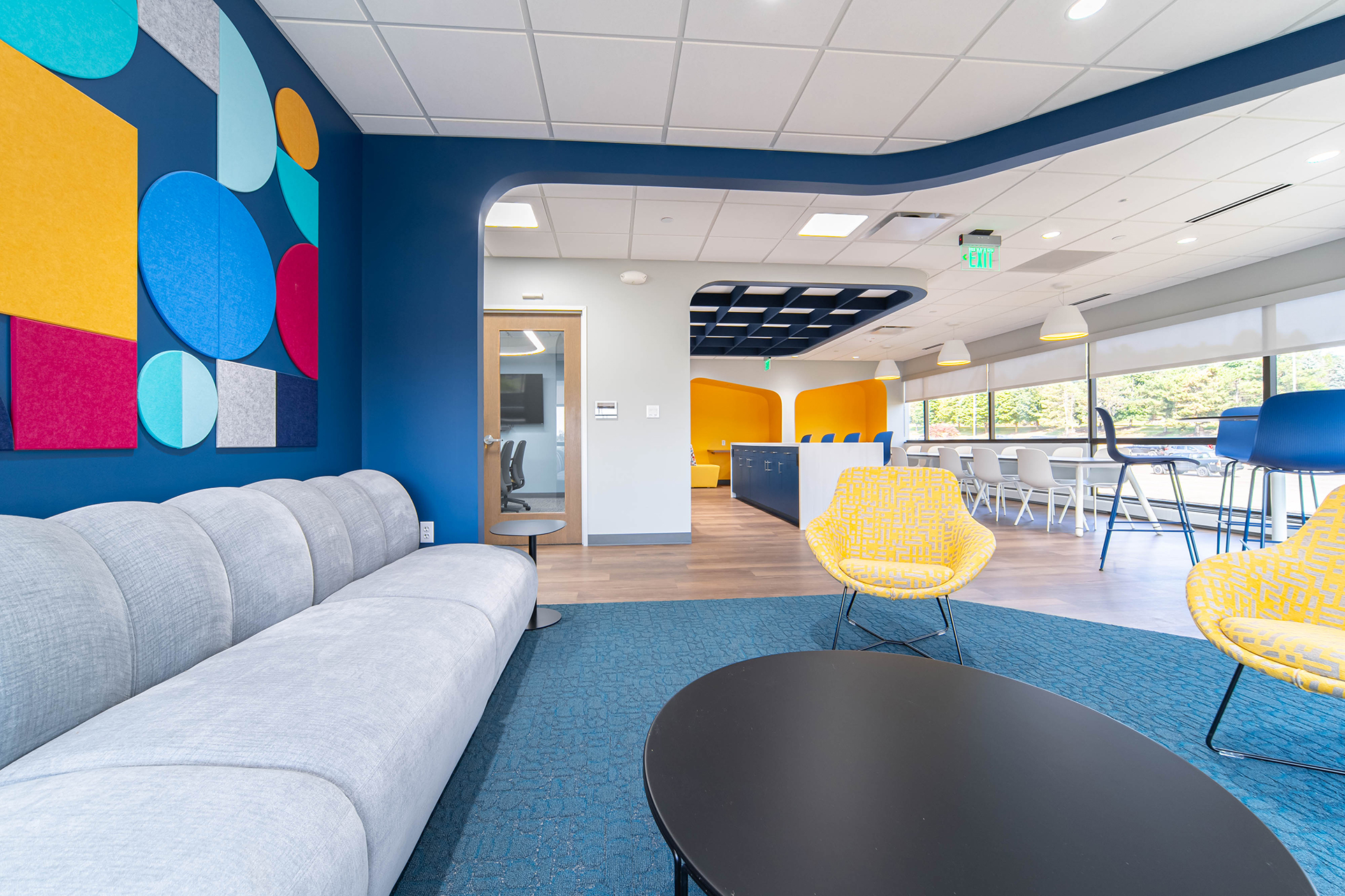 A modern office lounge features a gray sofa, round black table, and yellow chairs under an acoustic felt grid. Colorful geometric wall art adorns the left wall. In the background, a dining area with high tables and chairs enjoys natural light from large windows with chic pendant lights. .