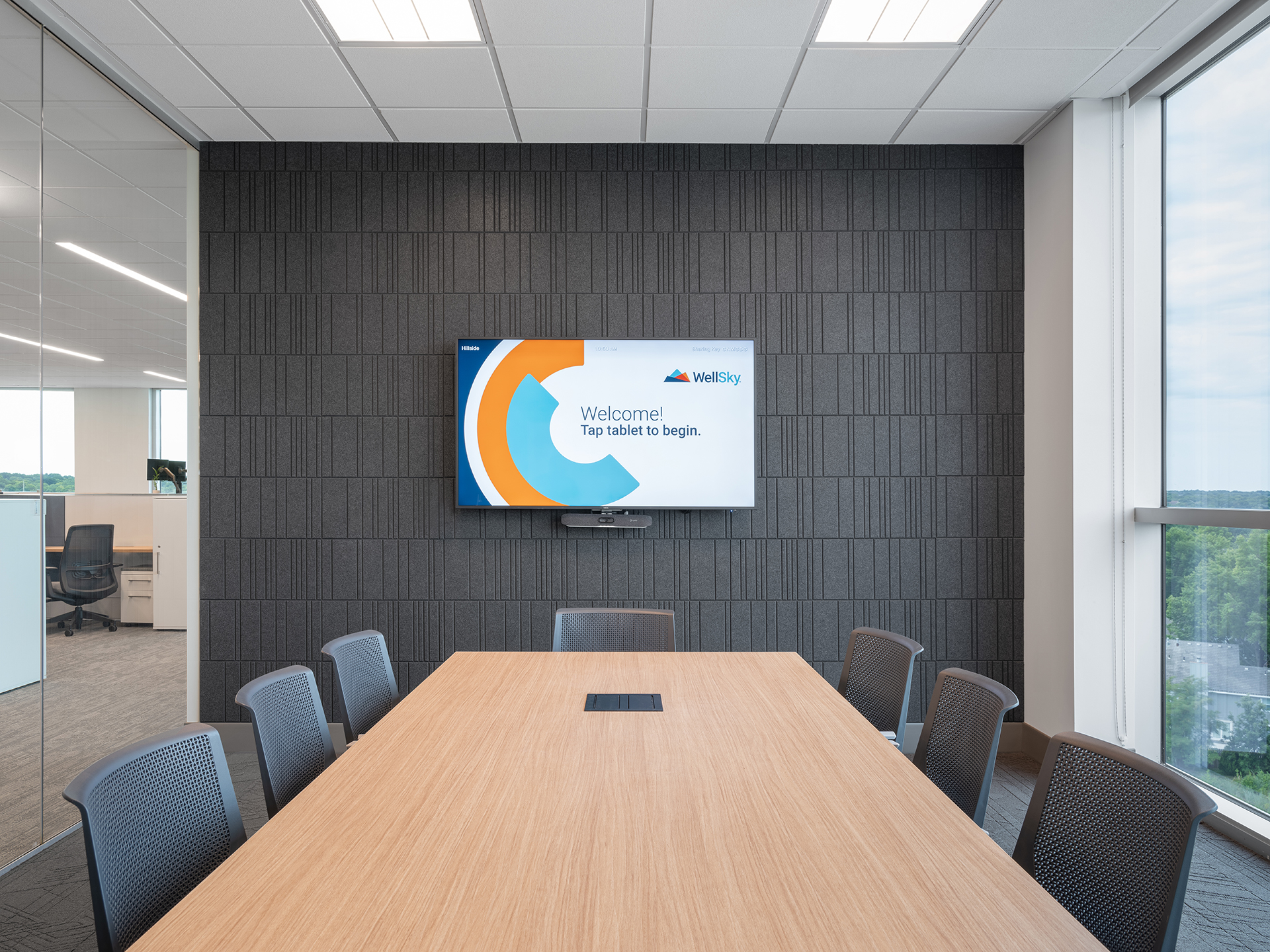 A modern conference room features a long wooden table surrounded by six black chairs. A flat-screen TV on the acoustic felt wall displays an interface reading, "Welcome! Tap tablet to begin." Well-placed windows flood the space with natural light from the right, enhancing the room's sleek design.