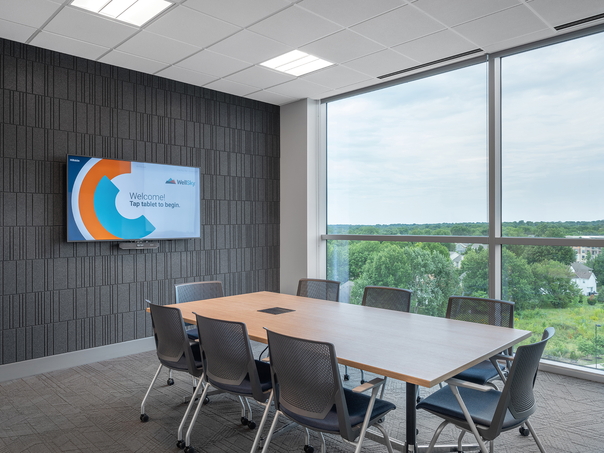 A modern conference room features a wooden table and six chairs. A large window offers a view of the verdant sky outside. The wall-mounted screen displays a welcome message, while walls boast textured barcode acoustic felt patterns. Ceiling lights are evenly spaced for optimal illumination.