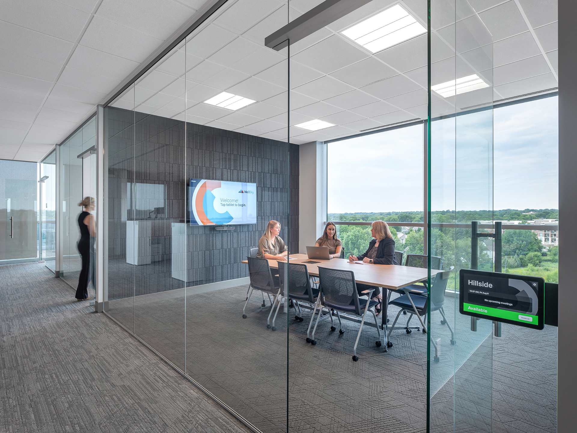 A modern office meeting room with sleek glass walls and polished acoustic felt wall tiles. Three people sit at a table with laptops in front of a large screen displaying a presentation. A person walks outside the room. A window offers a view of greenery and a well-lit skyline beyond the buildings.