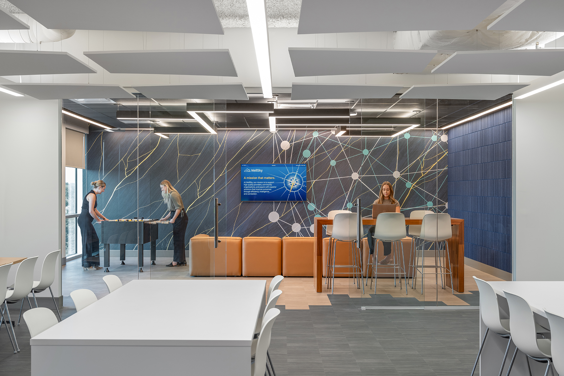 A modern office space with two people playing foosball and one person sitting at a high table with a laptop. The room features sleek furniture, a large display screen, and decorative acoustic felt wall tiles with geometric patterns that beautifully fracture the light.