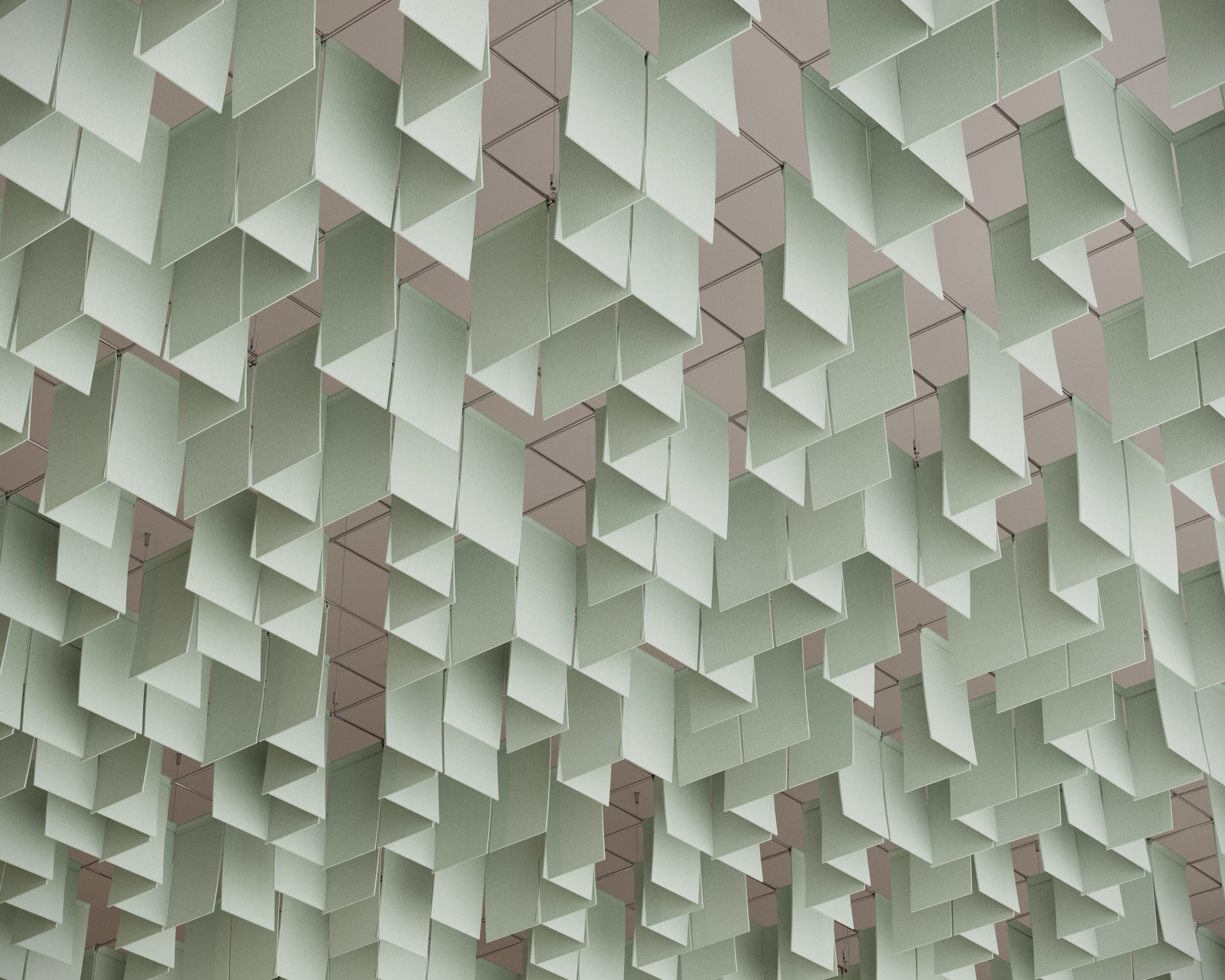 A ceiling art installation features numerous rectangular panels of pale green acoustic felt, suspended at various angles. These panels create a geometric pattern resembling floating clouds, with shadows accentuating the design against the neutral white ceiling backdrop.