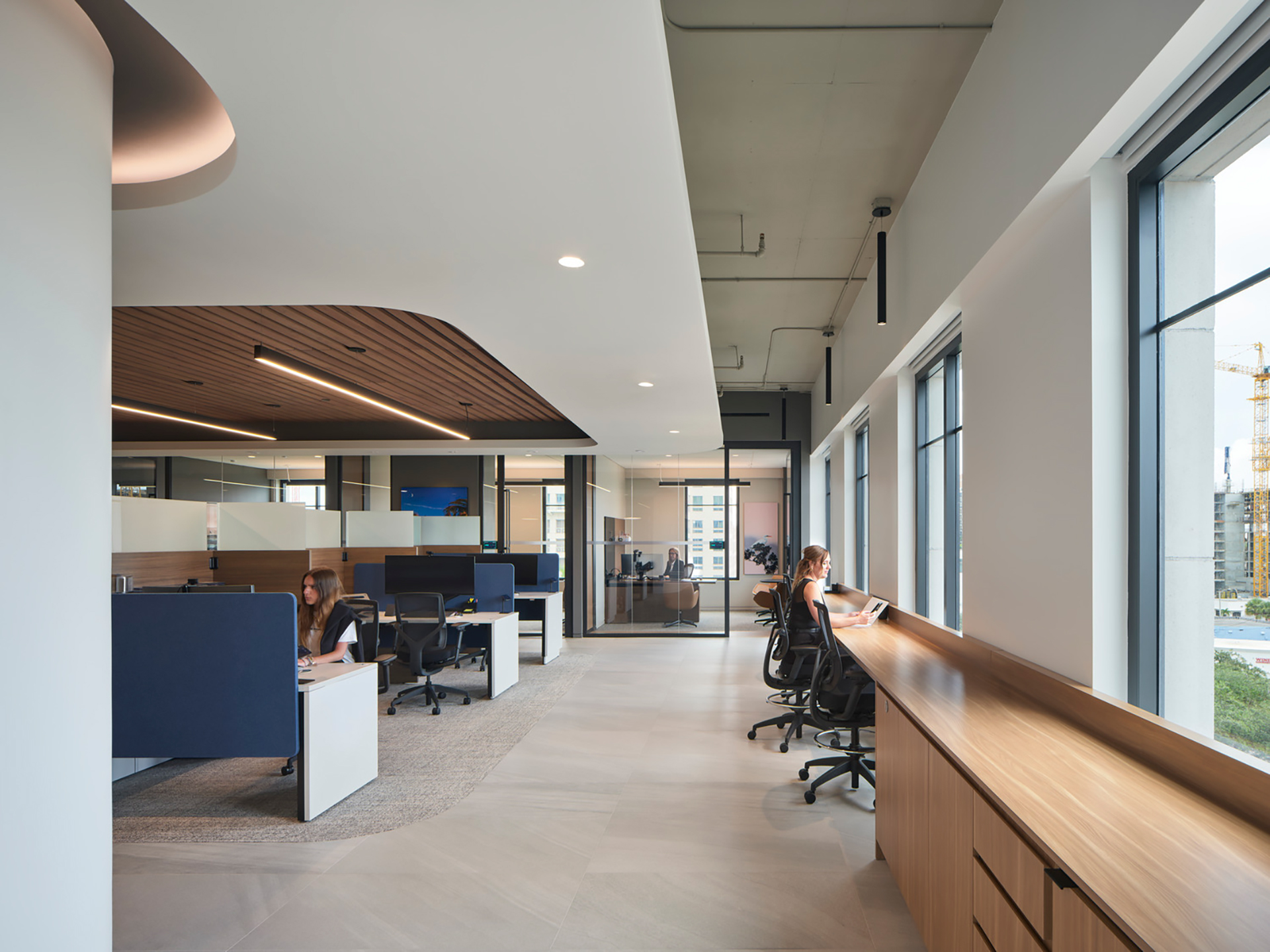 The modern office interior boasts large windows and abundant natural light. Employees work at desks and a long wooden counter beneath high ceilings. The open layout features partitioned workspaces, light wood accents, and an acoustic felt ceiling frame. A stunning cityscape is visible through the windows.