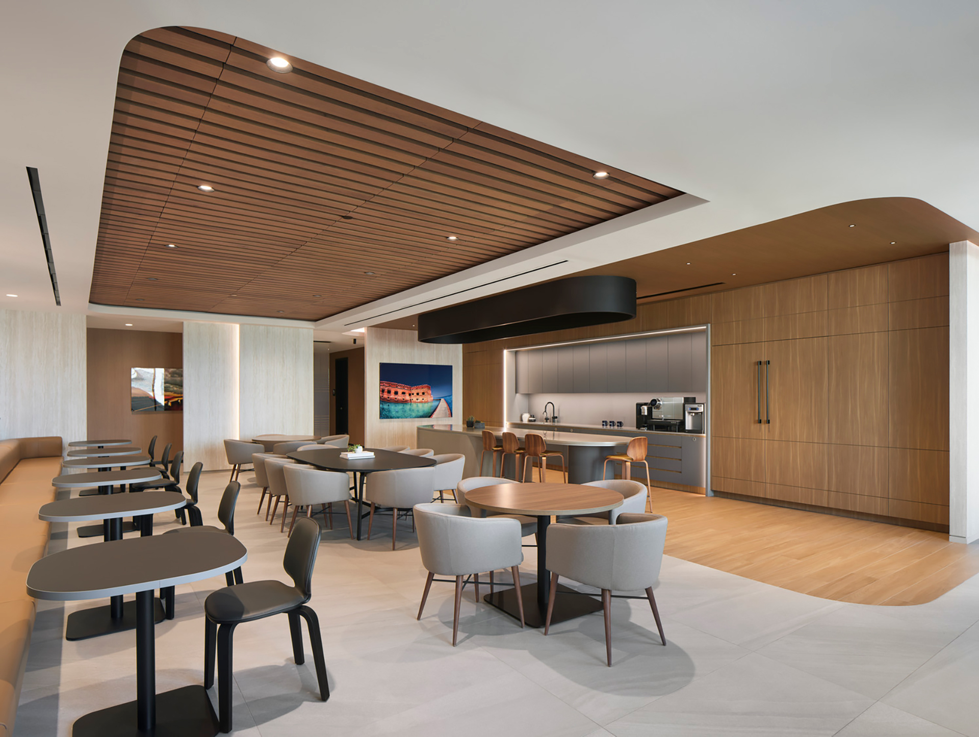 Modern open office lounge with a woodgrain acoustic felt ceiling frame, sleek black tables, and beige chairs. A kitchenette with coffee machines is in the corner, and a colorful painting adorns the light gray wall, subtly softening up the space.