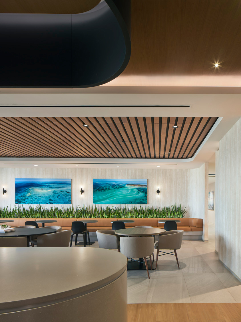 A modern interior featuring a seating area with round tables and cushioned chairs, softened by a long planter with lush foliage. Ocean-themed artworks line the back wall while a woodgrain ceiling frame enhances the space, adding warmth to its neutral tones.