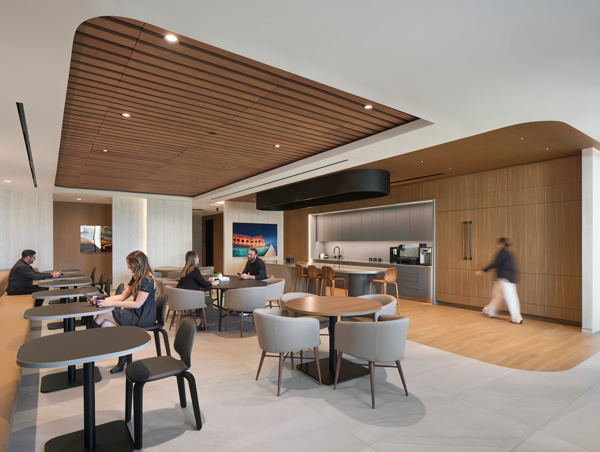 Modern office lounge with woodgrain acoustic felt ceiling frames, round tables, and gray chairs. People are sitting, using laptops. A kitchenette with a coffee machine is in the background. A person walks through, adding movement to the calm space.