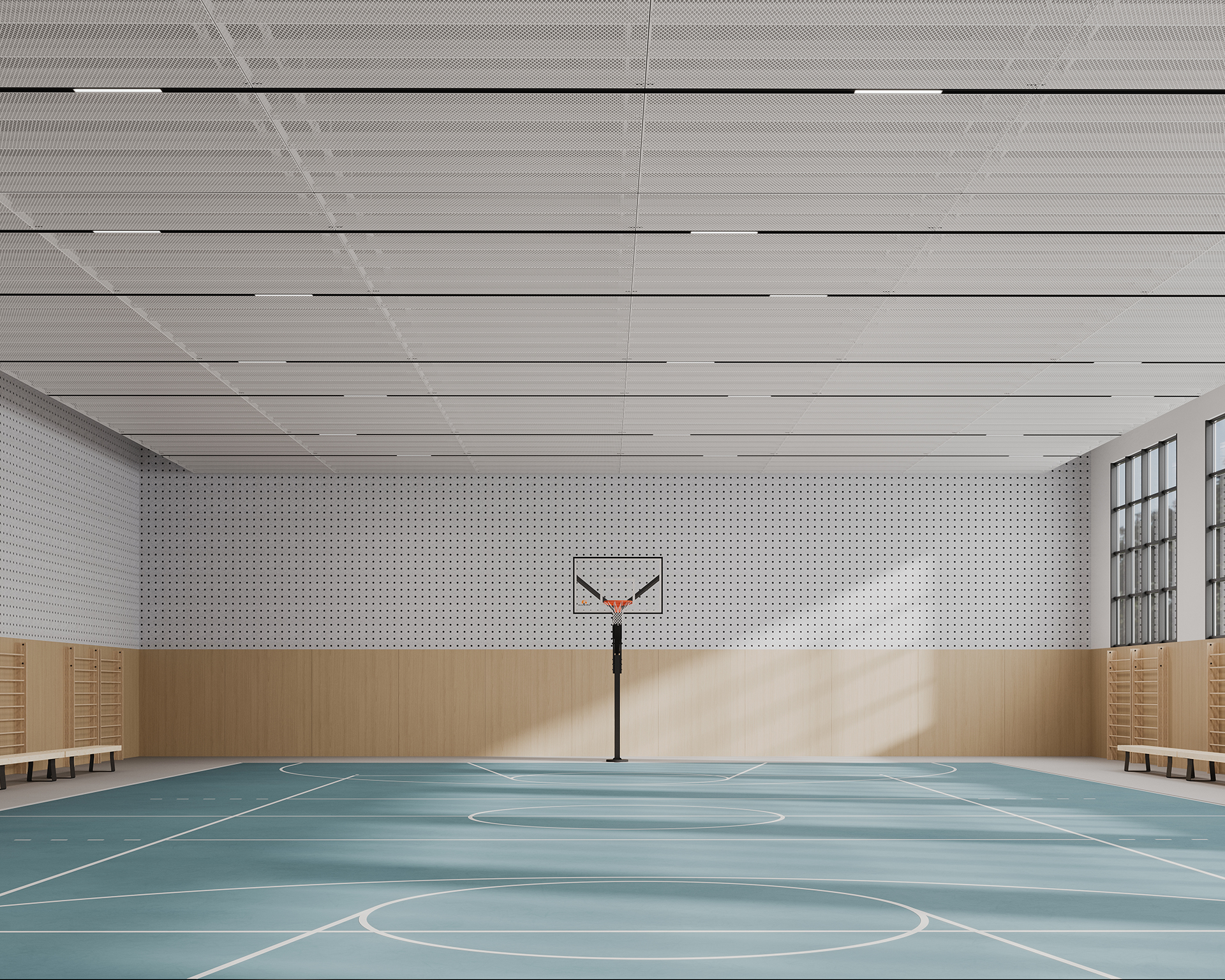 An empty indoor basketball court with a blue floor, featuring white lines marking the boundaries. A basketball hoop is positioned at one end. The space has a wooden lower wall, acoustic felt panels with small circle cutouts on the upper wall, and large windows on the right, letting in natural light.
