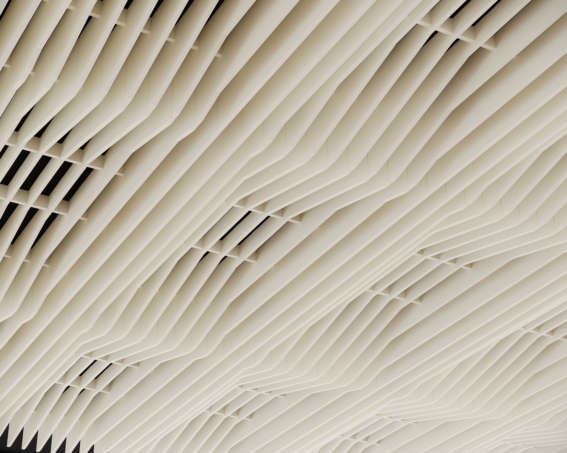 A close-up view of an acoustic felt ceiling with a geometric pattern featuring parallel, wavy lines and vertical slats. The design creates a visually striking texture and depth. The color scheme is a light beige or off-white.