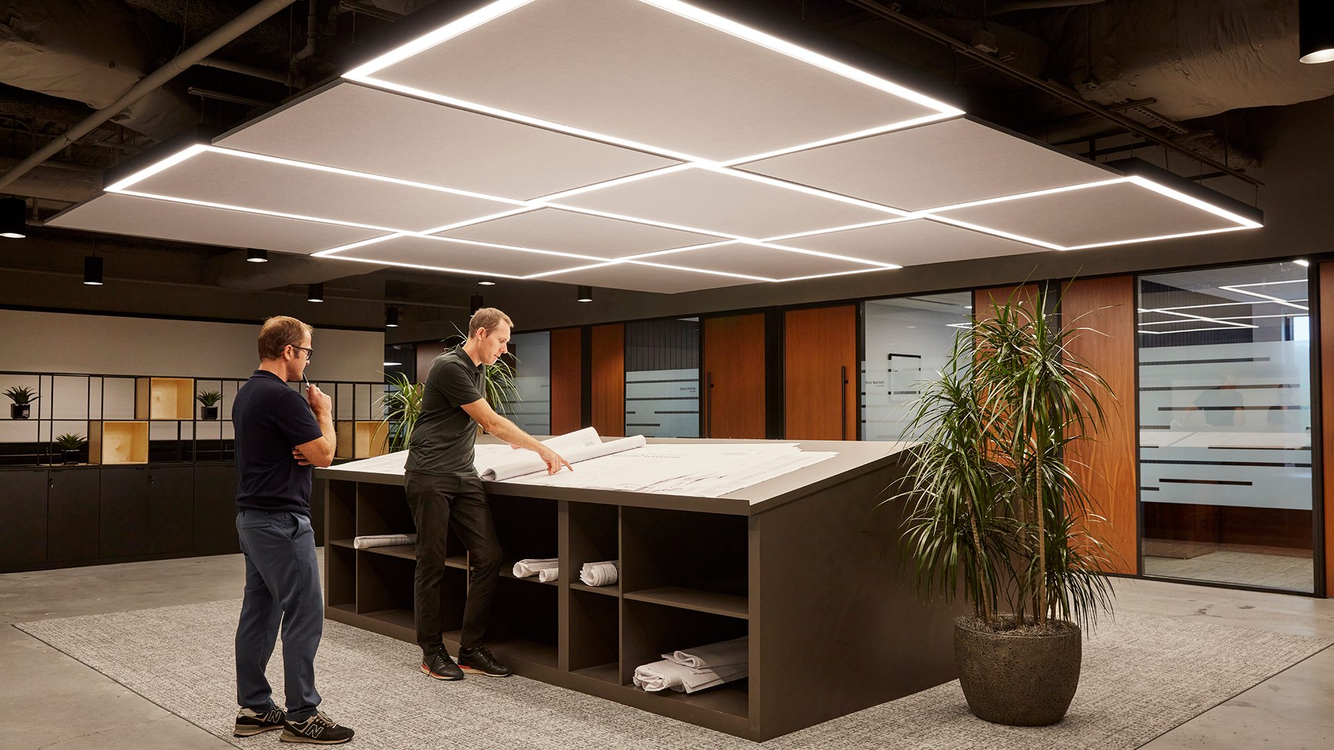 Two men stand at a large, slanted workspace reviewing architectural plans in a modern office. One wears a black shirt, and the other a blue shirt. The room is well-lit with geometric ceiling lights around acoustic felt ceiling panels and features large glass partitions and potted plants.