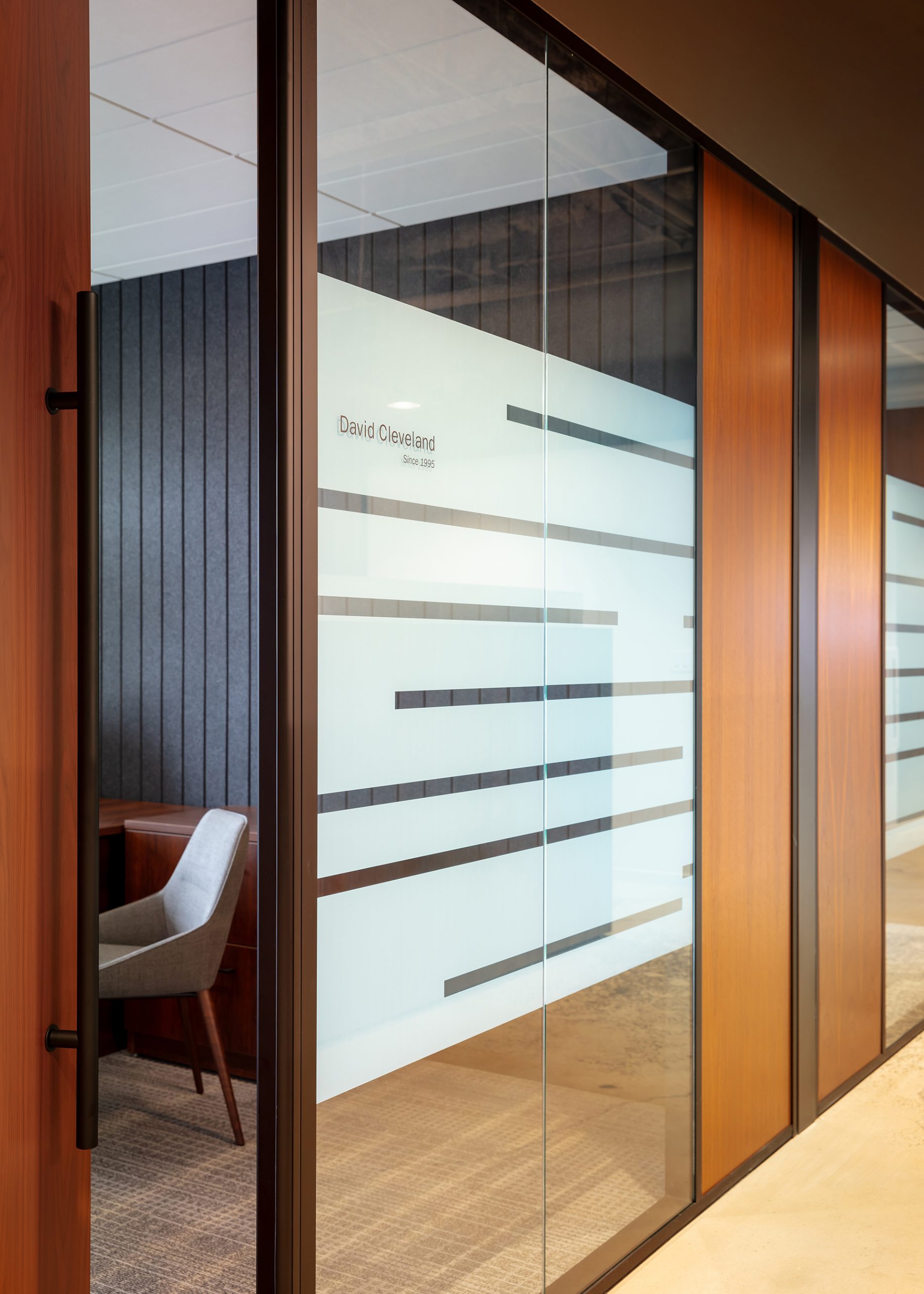 Glass office doors with wooden frames have horizontal frosted lines. A sign reads "David Cleveland." Inside, a gray cushioned chair and a wooden desk are visible against a dark paneled acoustic felt wall.