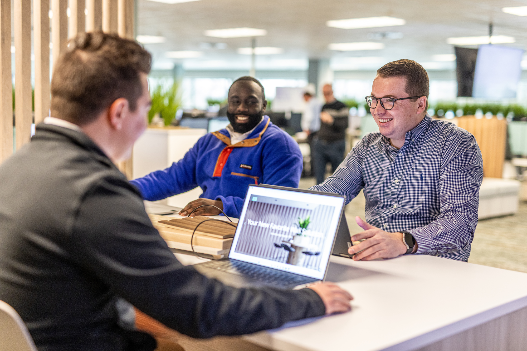 Three people sit at a table in a modern office, two facing one with a laptop open, displaying an Acoufelt website. They are smiling and appear engaged in discussion. The background is bright with plants and workstations, reflecting an atmosphere of team growth.