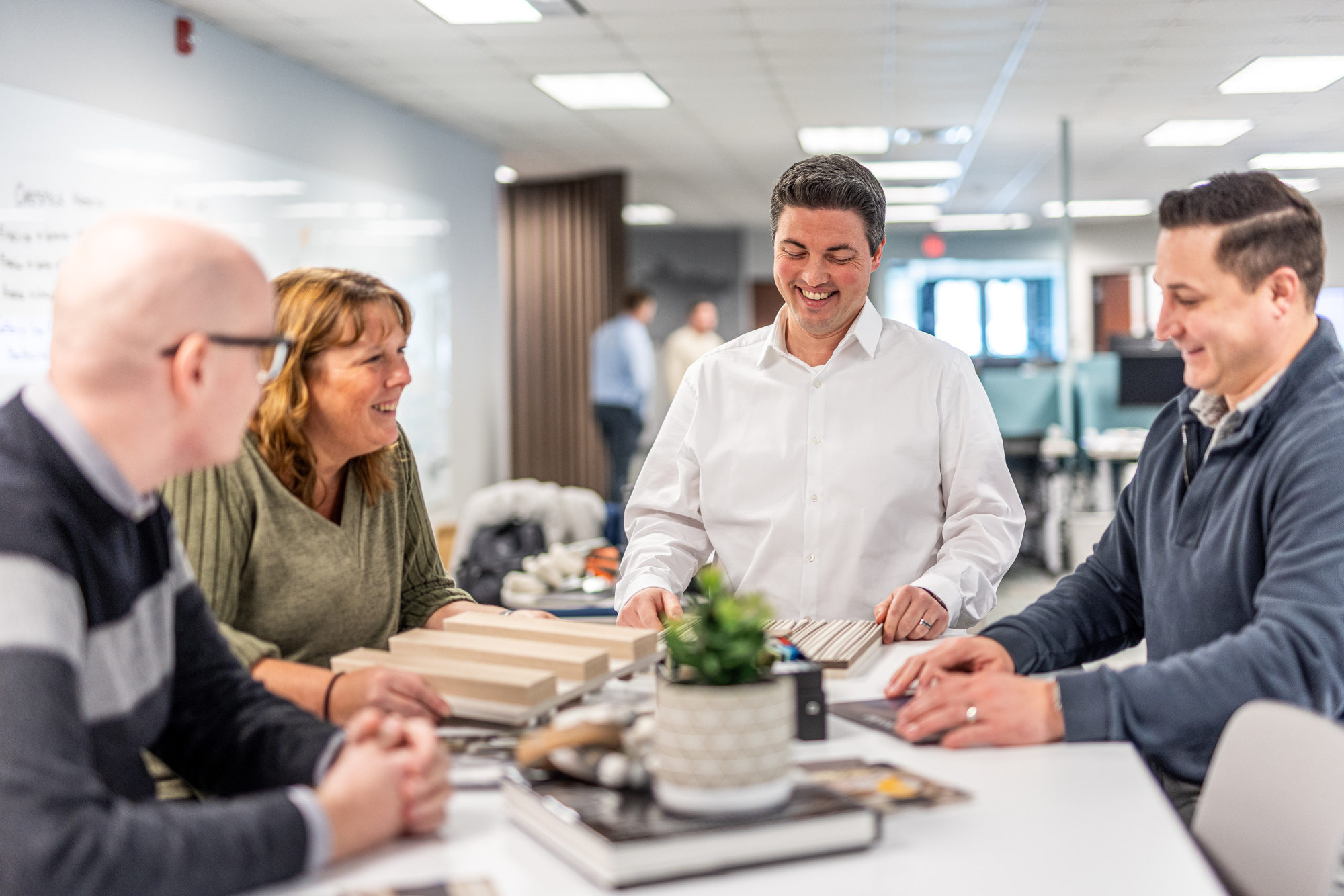 A team of four people gathers around a table in a bright office, smiling as they explore Acoufelt samples. A potted plant and open notebooks add to the casual, collaborative atmosphere, embodying a spirit of future-focused growth.