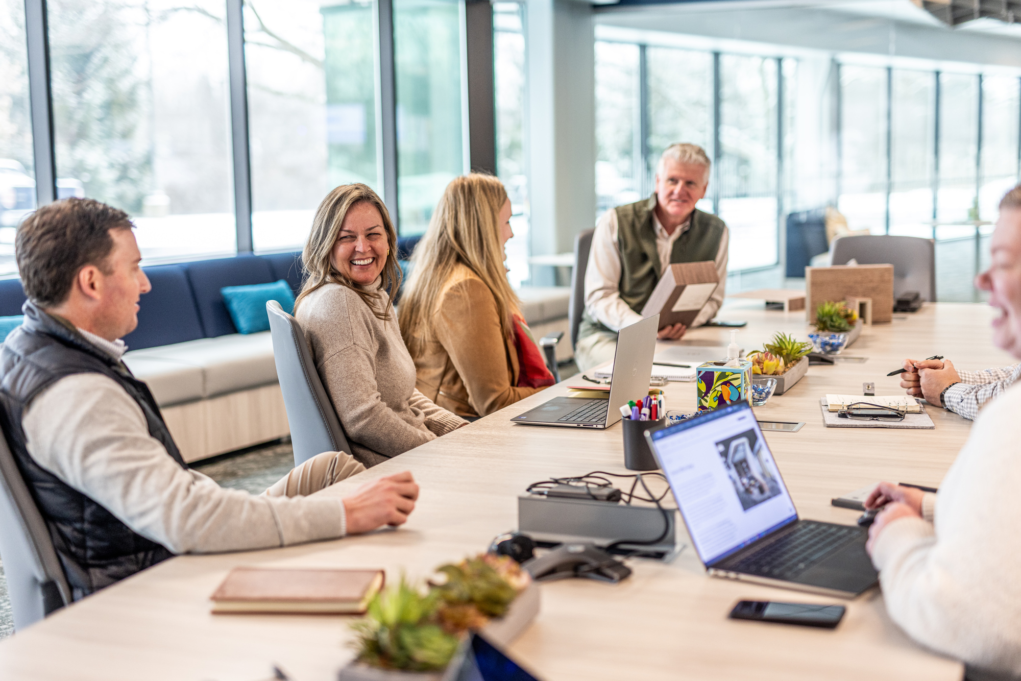 A group of people sitting at a conference table in an office, engaging in a discussion about Team Growth. Laptops, notepads, and small potted plants are on the table. Large windows in the background provide natural light. Everyone appears to be smiling or attentive.