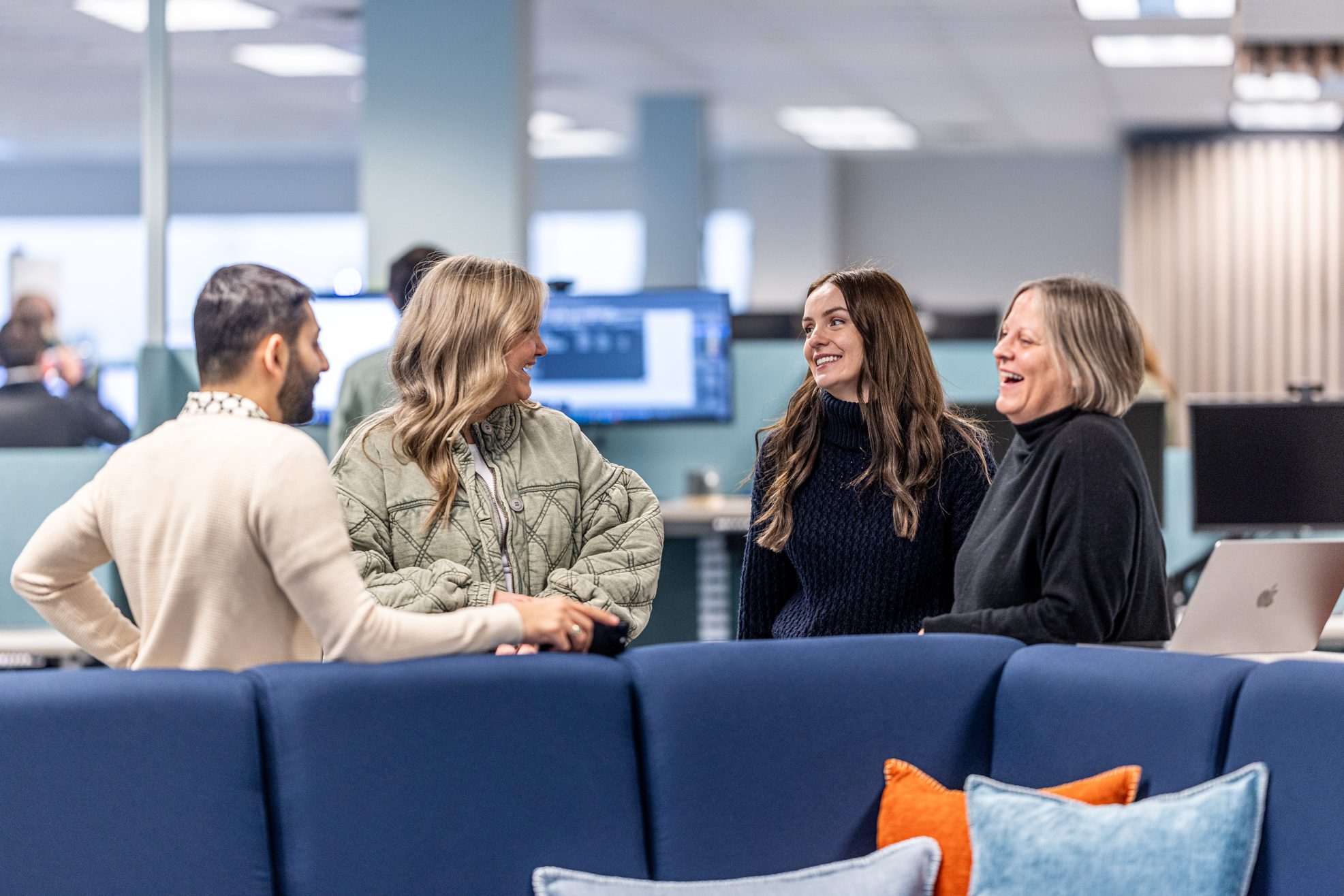 Four people are standing and talking in an office with computers in the background, embodying the spirit of team growth. They appear to be engaged in a friendly conversation, with two women smiling widely. Blue cushions are visible on a sofa in the foreground, setting a cozy tone for building the future together.