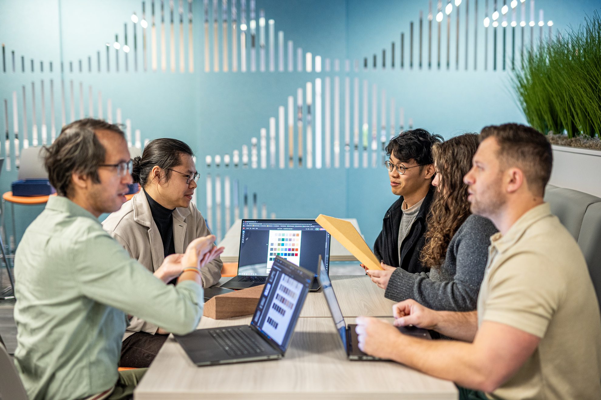 Five people sit around a table in a modern office, laptops open as they collaborate on building the future. Surrounded by green plants, this diverse group in casual attire is focused on an Acoufelt presentation onscreen, embodying team growth in action.