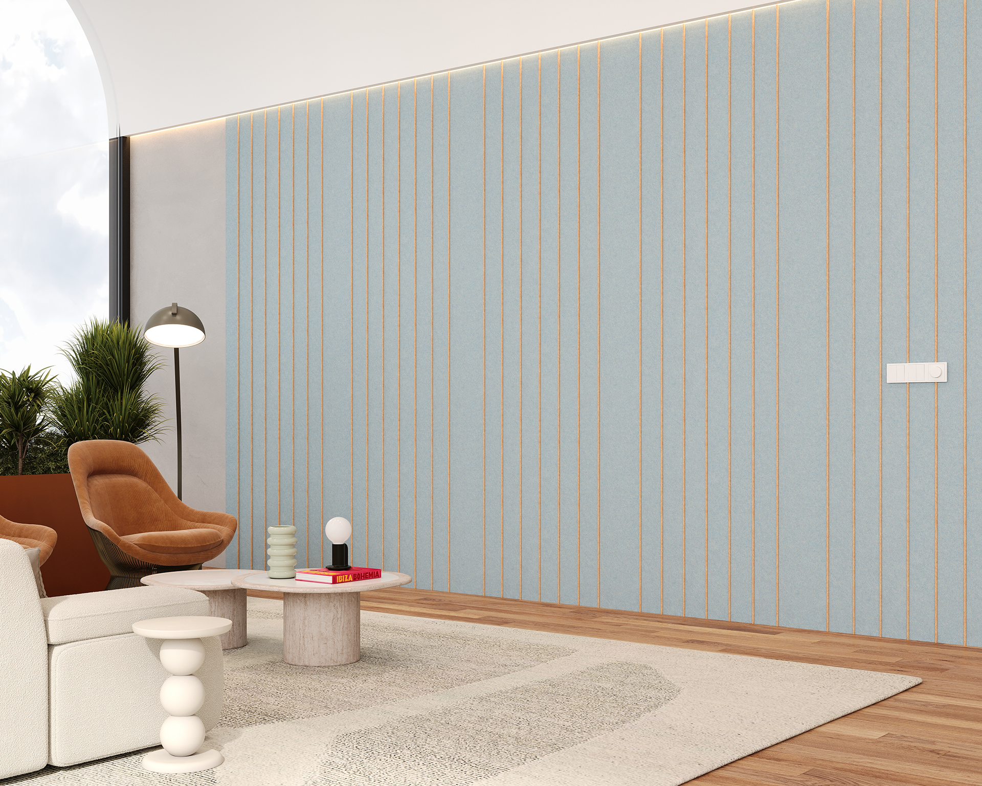 Modern living room with a blue and gold vertically striped acoustic felt accent wall. It features a white sofa, brown armchair, round coffee table, and a floor lamp. There are green plants near a large window, and the floor is light wood with a patterned rug.