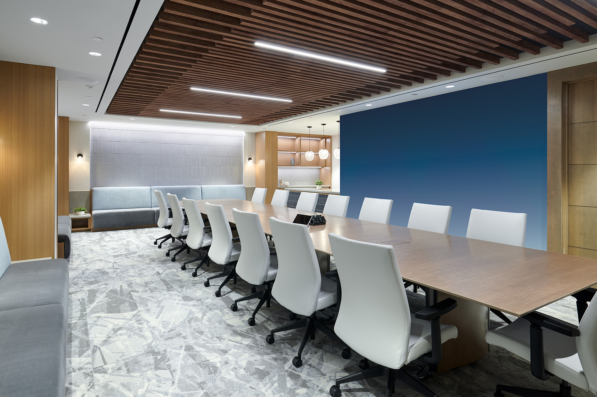 Modern conference room with a long wooden table surrounded by white office chairs. The room features a woodgrain slat acoustic felt ceiling, blue accent wall, and carpeted floor. Shelves with decorative items are visible in the background.