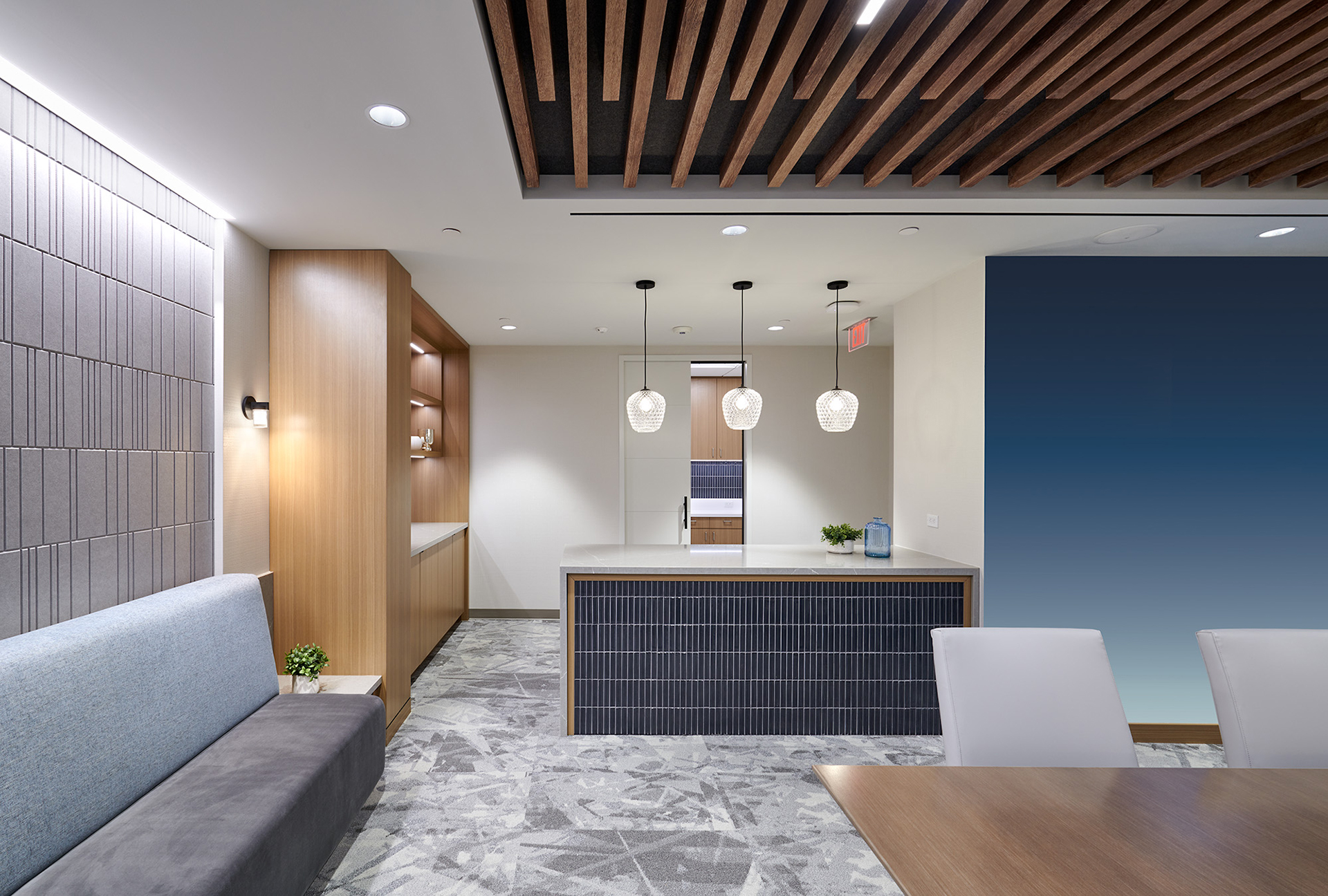 Modern office lounge with woodgrain acoustic felt ceiling panels and a mix of soft gray and wood tones. Features a seating nook on the left, a central bar with three pendant lights, and an acoustic felt accent wall on the right. Geometric-patterned floor tiles add detail.