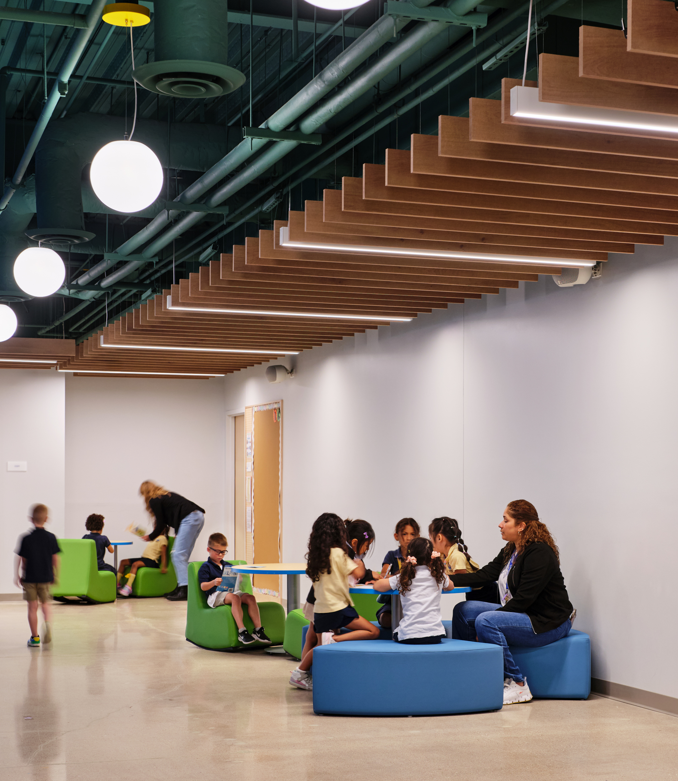 Children and adults sit and talk in a modern, brightly lit hallway at Magellan International School, featuring acoustic felt ceiling baffles, circular seats, and decorative panels. Some kids gather at tables while others move around in the background.