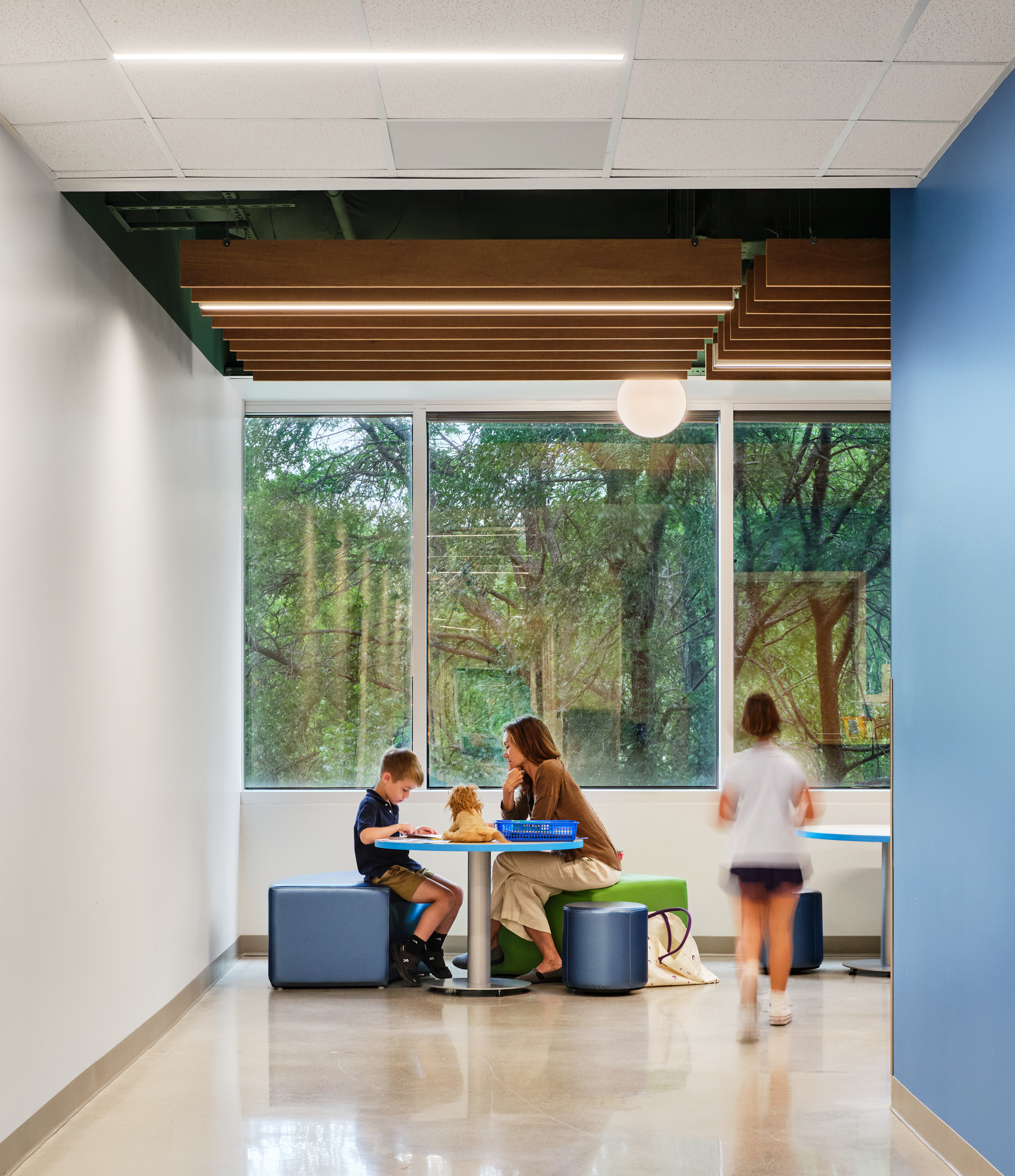 A woman sits at a table with a young boy, reading a book, while a child walks by in a bright hallway at Magellan International School, featuring large windows and acoustic felt ceiling baffles above.