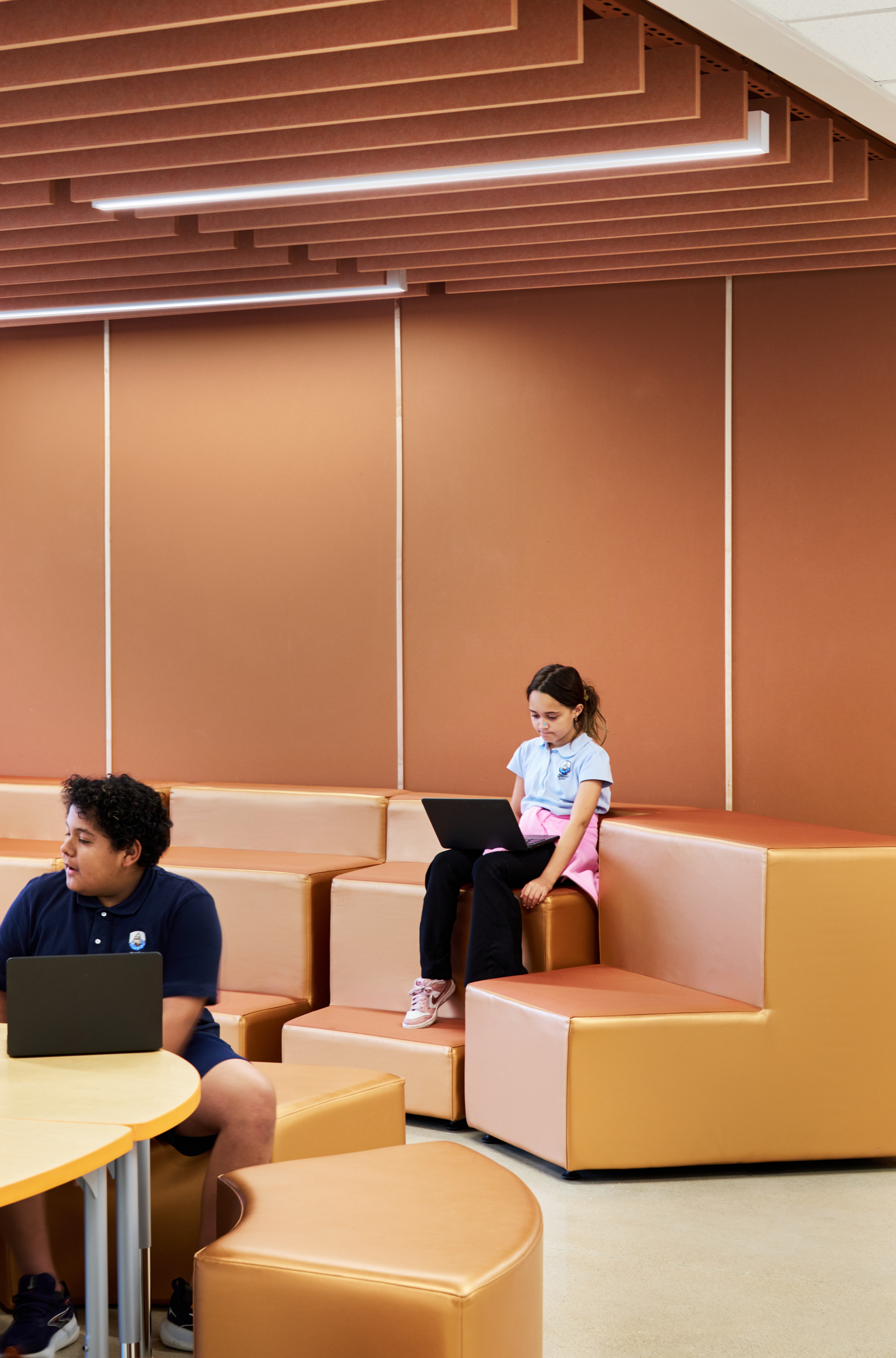 A young girl sits on tan, modular seating with a laptop, focused on her work. Another student sits nearby at a round table, also using a laptop. The space features brown walls and acoustic felt ceiling baffles.