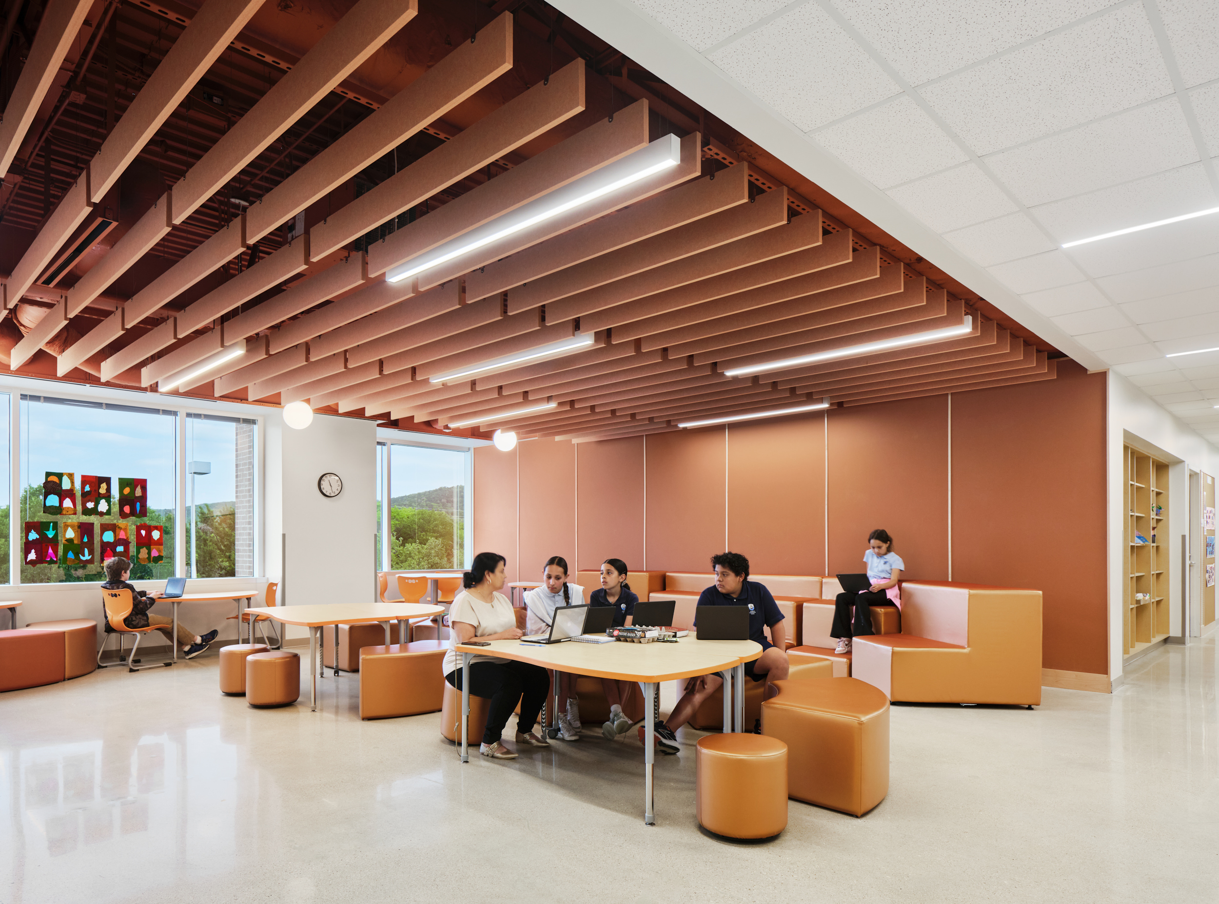 Five students sit and work together at a table in a modern, bright lounge with orange seating and distinctive acoustic felt ceiling baffles; one student sits alone on a bench, using a laptop.