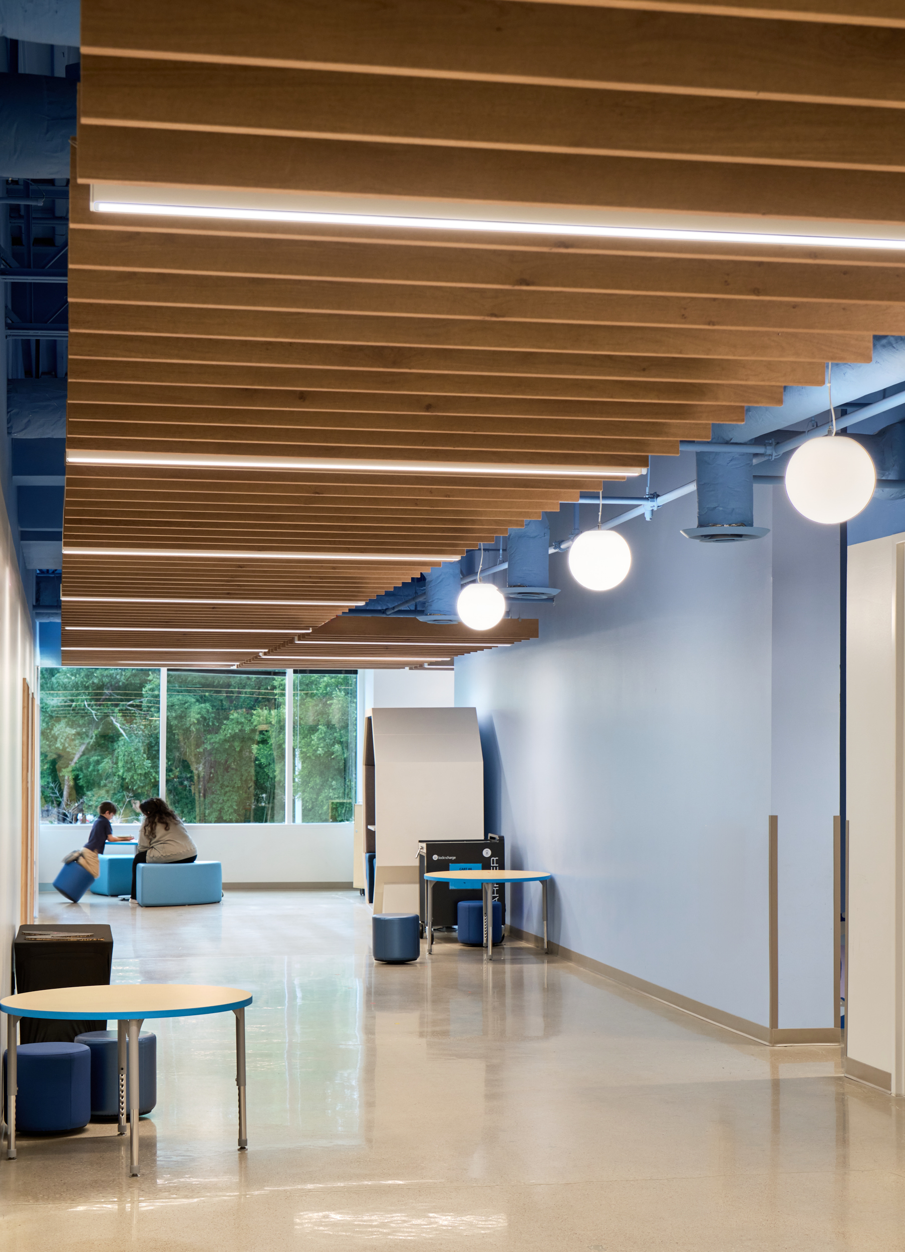 A modern hallway at Magellan International School features light blue walls, woodgrain acoustic felt ceiling beams, linear and arc ceiling baffles, round hanging lights, and large windows. Two people sit and talk near the window with desks and stools along the hallway.