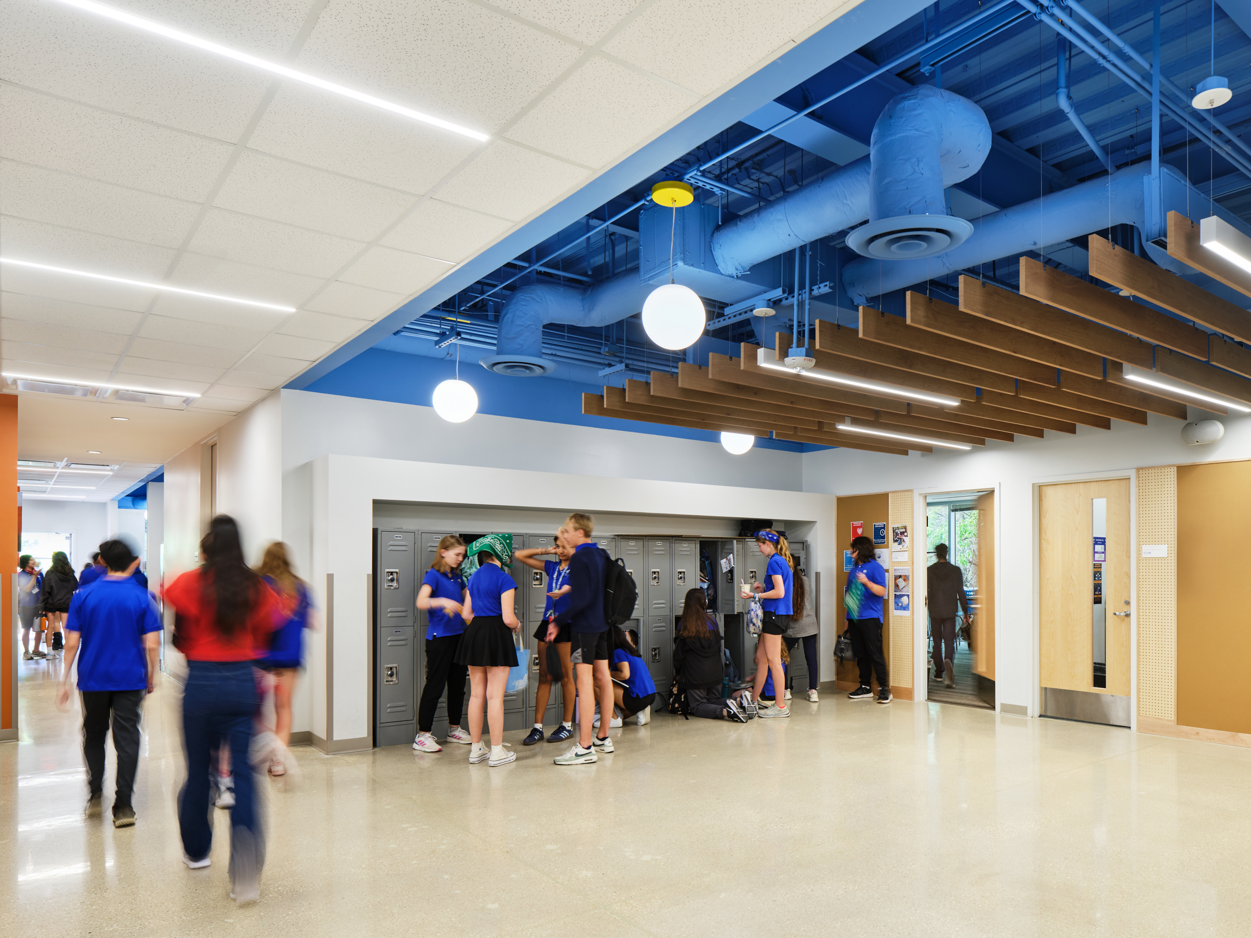 A group of students in blue uniforms gather and talk near lockers in a brightly-lit school hallway at Magellan International School, featuring modern design elements like acoustic felt ceiling baffles, exposed blue pipes, and wooden ceiling slats.