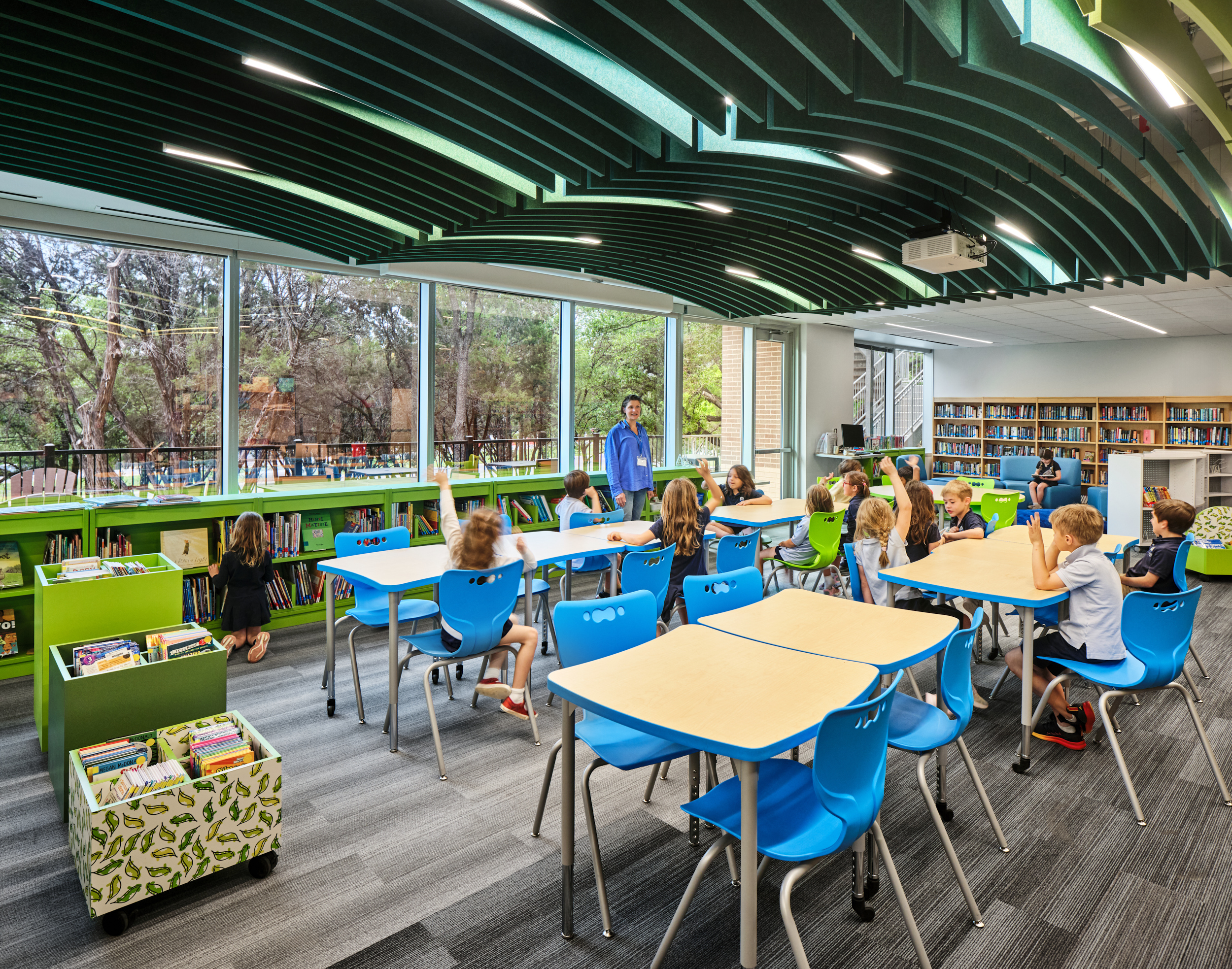 Children sit at tables in a brightly lit Magellan International School library classroom with large windows. Arc acoustic felt ceiling baffles add style above, while blue chairs and green accents bring color to the space lined with bookshelves.