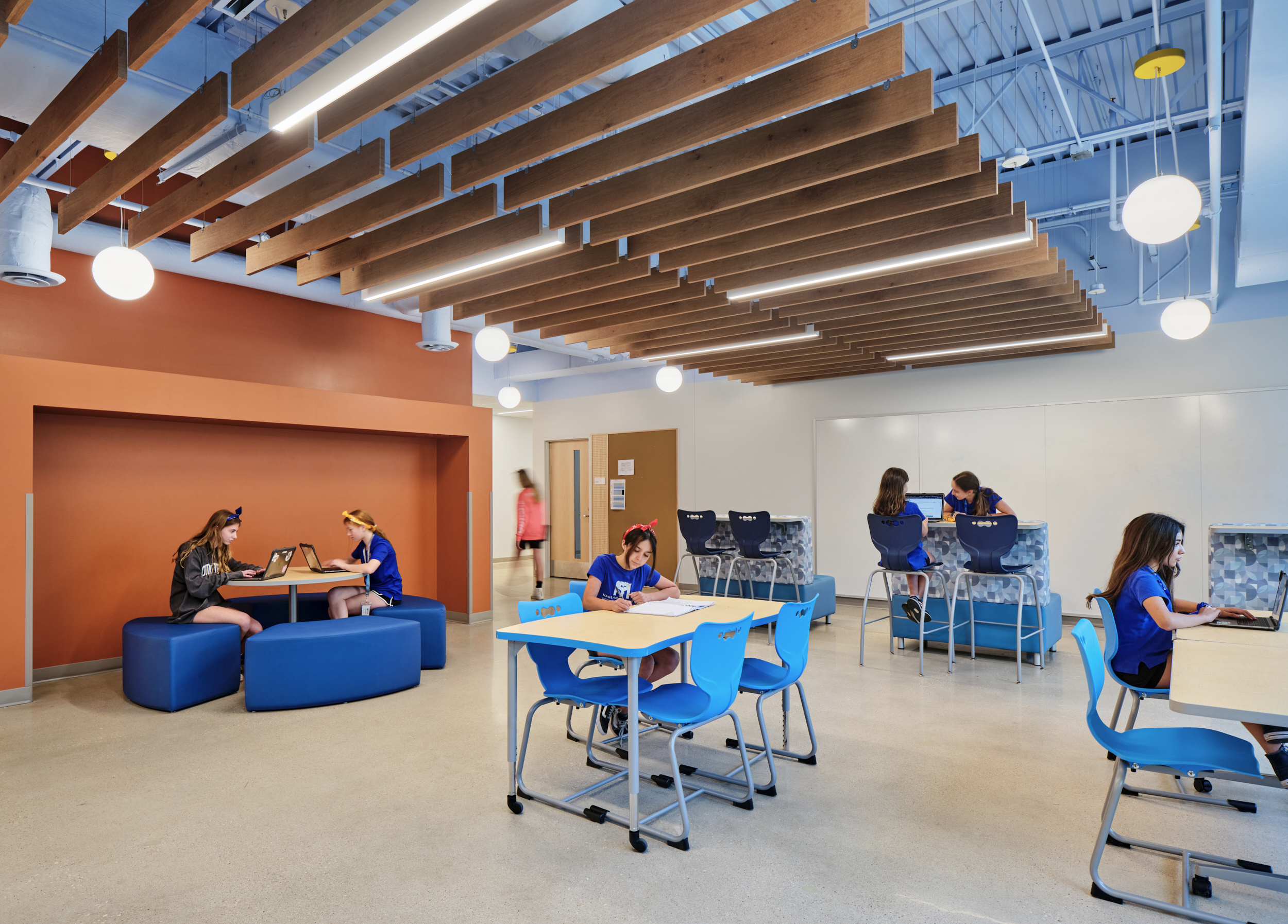 Students work and study in a modern classroom at Magellan International School featuring blue furniture, wood panels, and bright lighting enhanced by linear acoustic felt ceiling baffles. Some sit at tables, while others relax on cushioned benches in a cozy alcove.