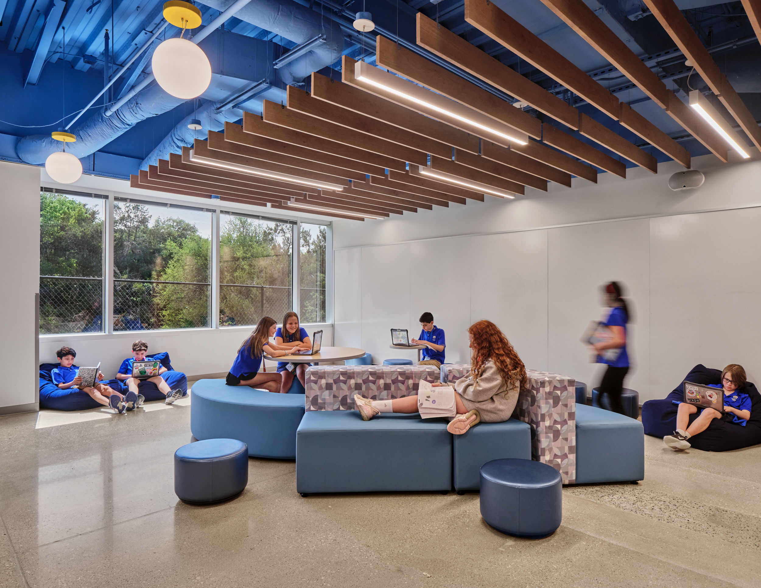 A group of students in blue shirts study, read, and use laptops on modern blue seating in a bright room at Magellan International School, featuring linear acoustic felt ceiling baffles, large windows, wooden beams, and polished concrete floors.